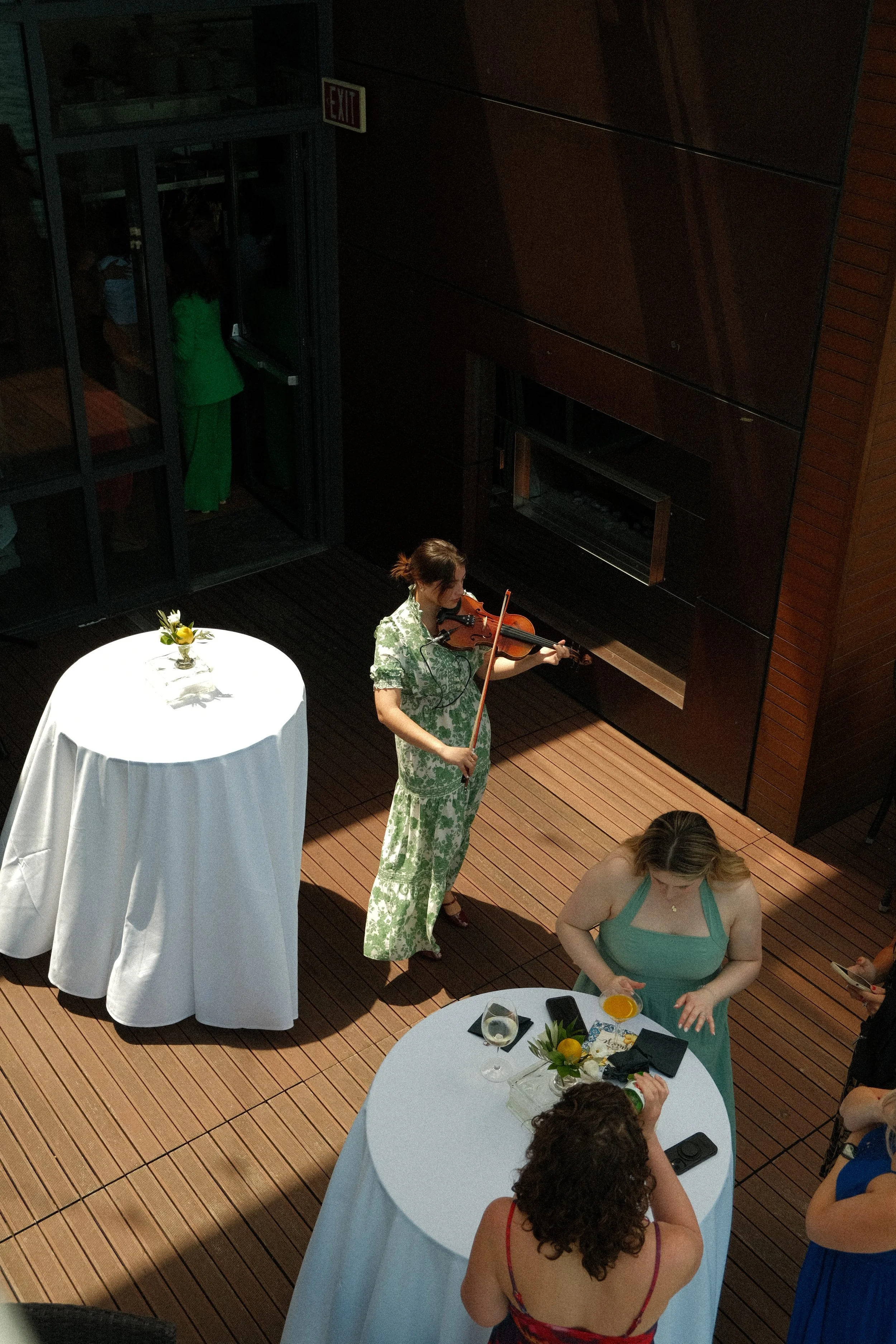 A woman playing the violin at a round table with drinks and a flower arrangement. Several other people are at nearby tables in a wood-paneled room with a dark wall and glass door. Some people are dressed in colorful attire.