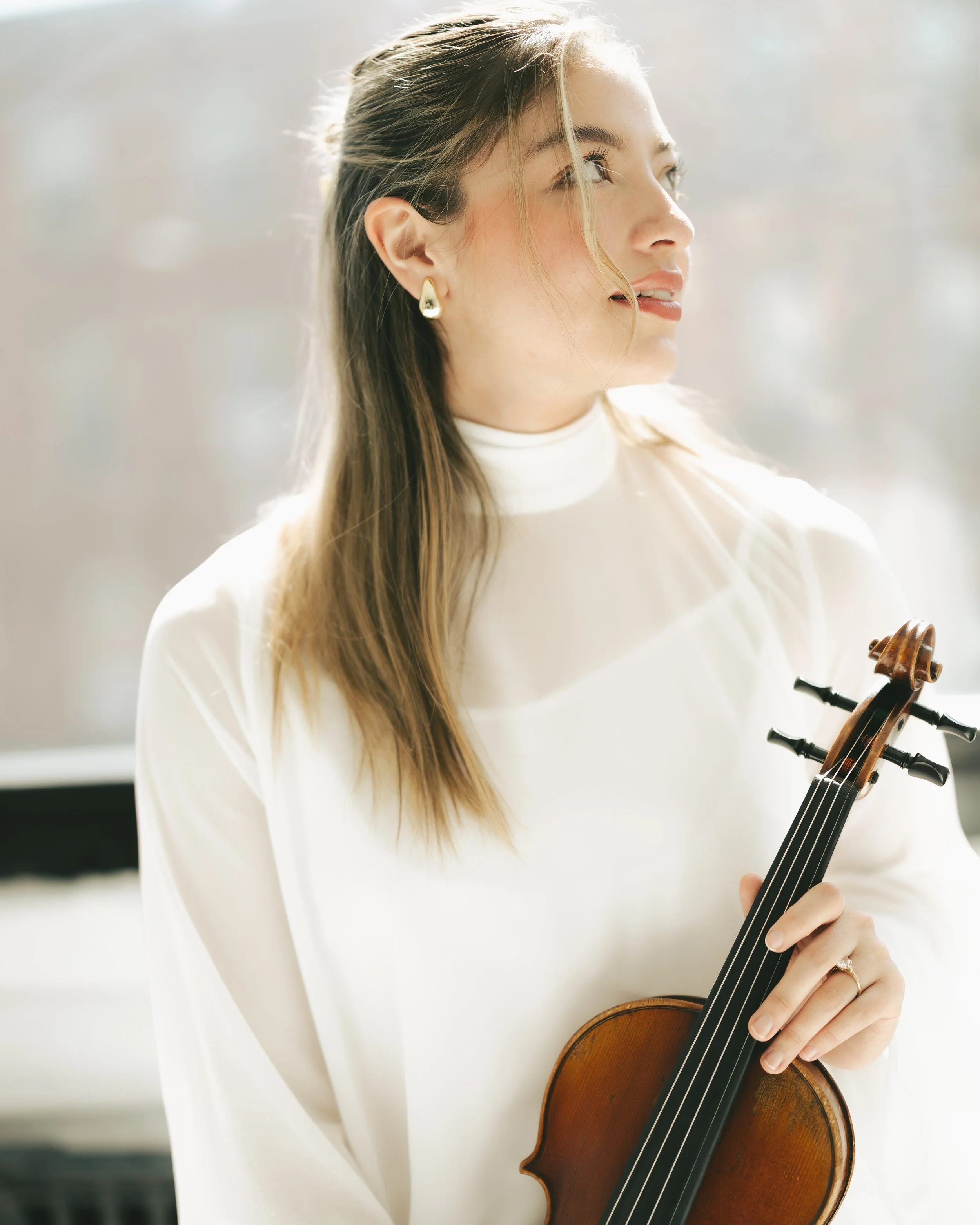 A woman wearing a white top plays the violin, with sunlight illuminating her brown hair and a soft, blurred background.