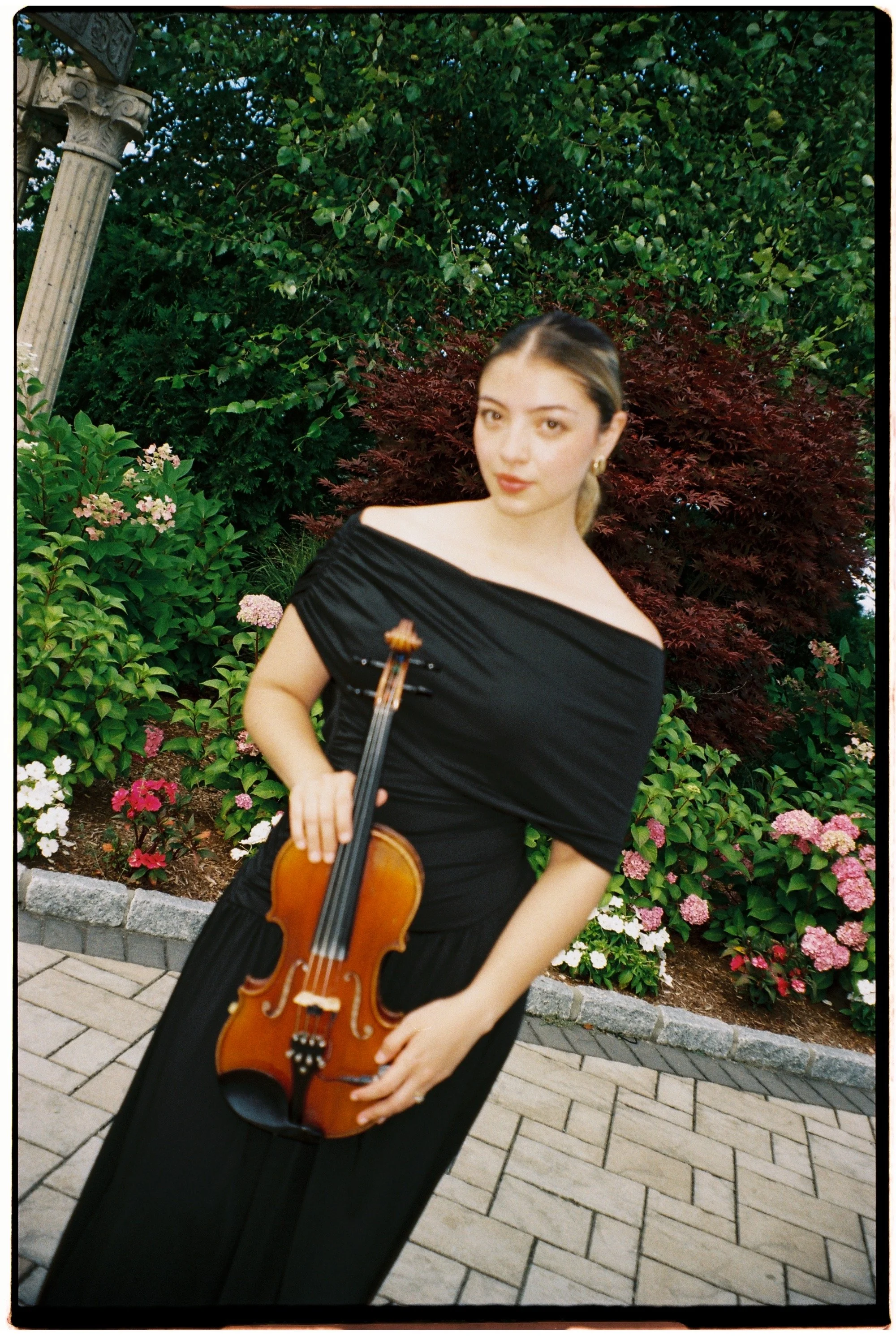 A woman in an elegant off-shoulder black dress holding a violin and bow, standing outdoors on a paved walkway with colourful flowering bushes and green trees in the background.