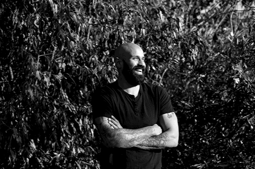 A man with a beard and tattoos, wearing a black shirt, standing in front of bushes, smiling and looking to the right.