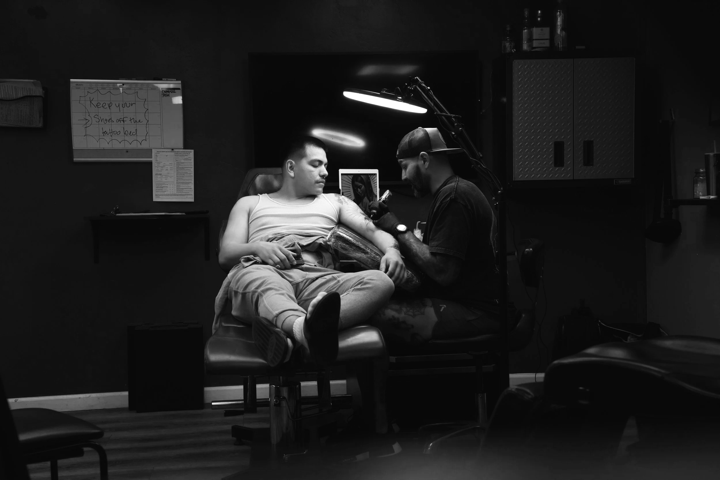 Black and white photo of a man receiving a tattoo on his arm from a tattoo artist in a tattoo studio. The man is sitting in a chair with his eyes closed, and the artist is wearing gloves and a cap, focused on the tattoo work.
