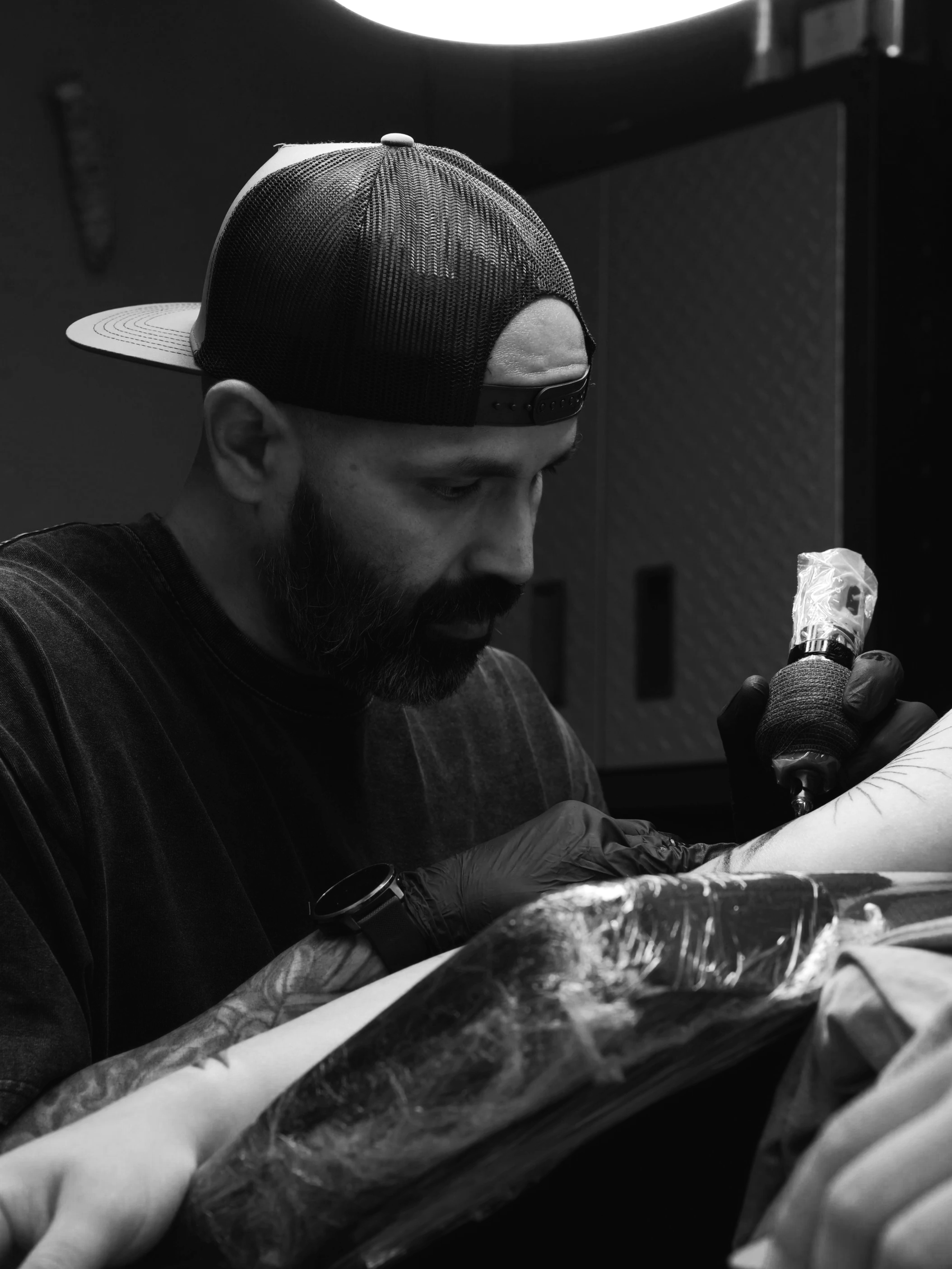 A man with a beard and cap tattooing a person's arm in a tattoo shop. The man is wearing gloves and is focused on his work.