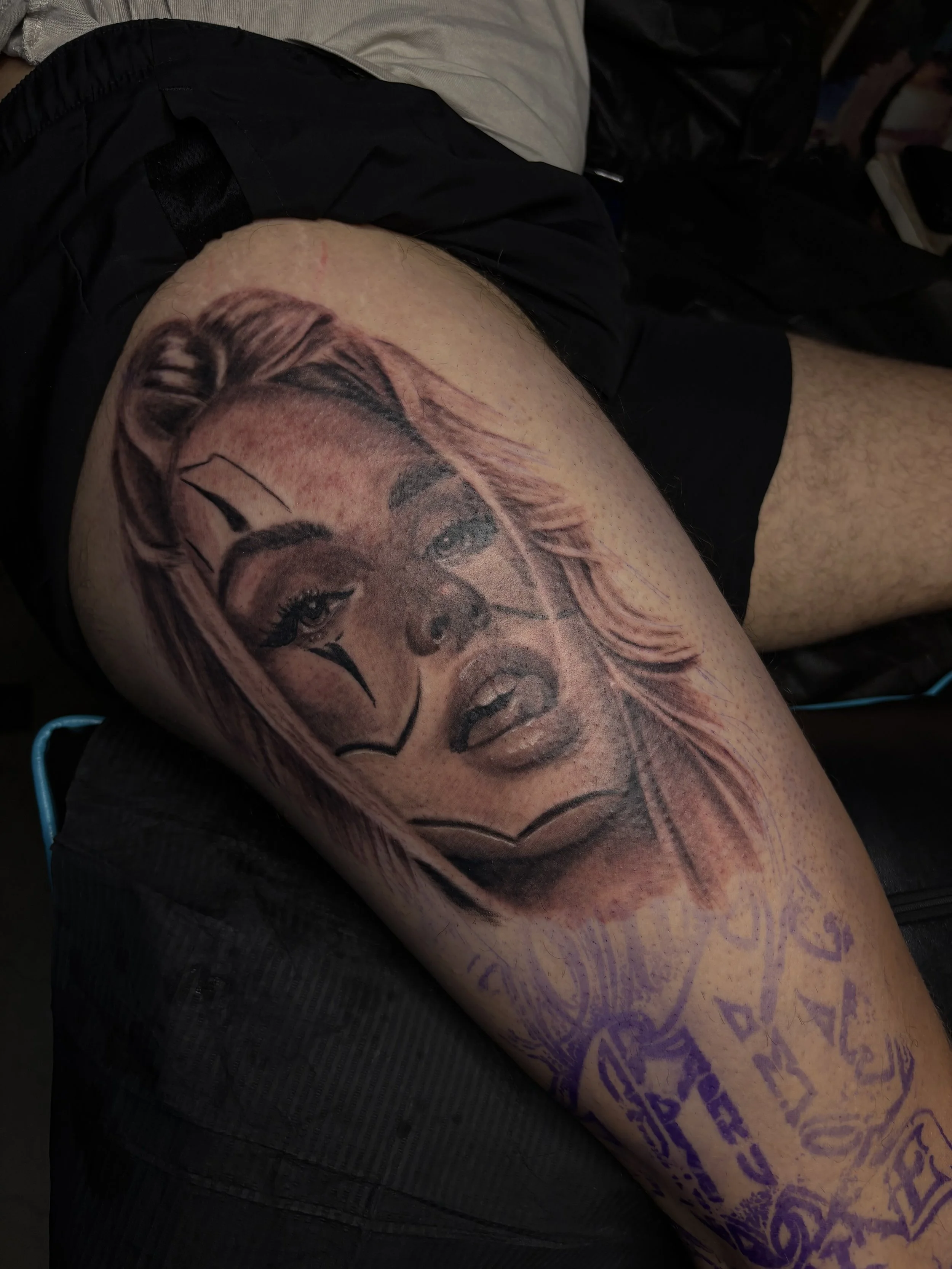 A person with a large, detailed portrait tattoo on their thigh, featuring a woman's face with styled hair and makeup, along with purple lettering.