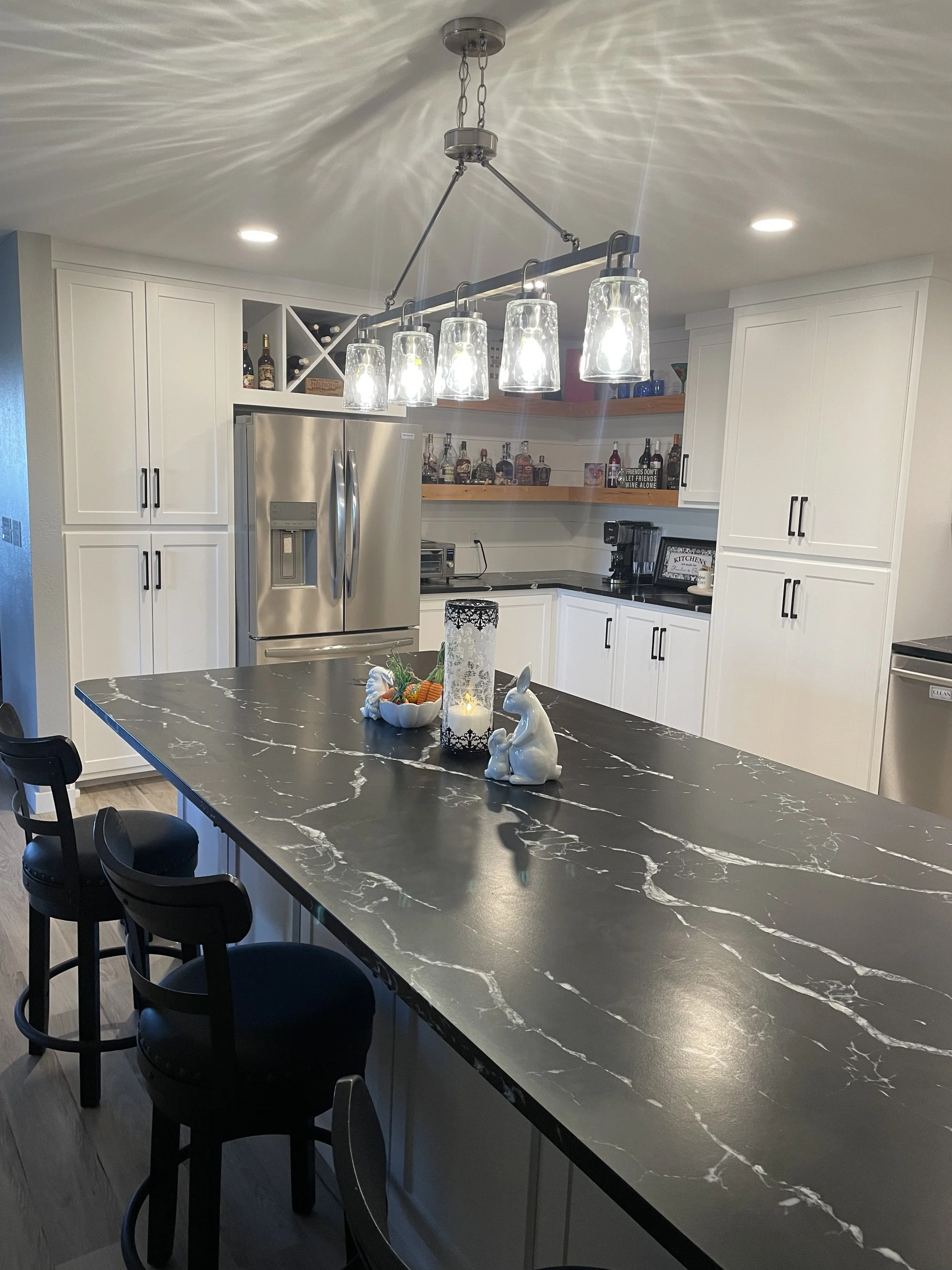 A modern kitchen with white cabinets, stainless steel refrigerator, black marble countertop island, and decorative bunny and carrot figures, lit by hanging glass pendant lights.
