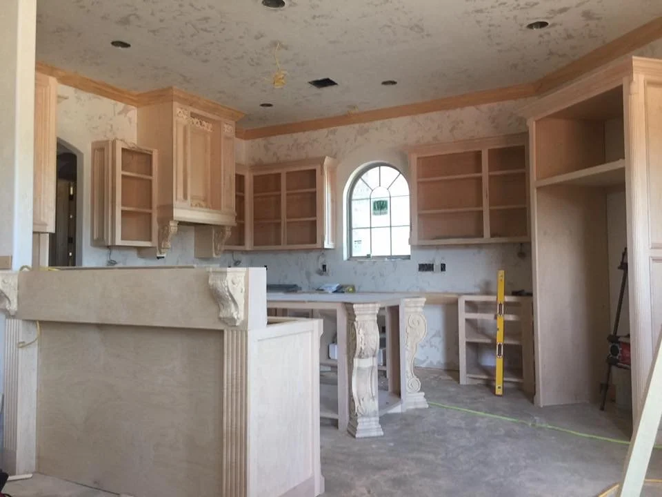 Kitchen under construction with unfinished wooden cabinets, some open shelves, and decorative columns supporting the countertop, with an arched window and construction tools visible.