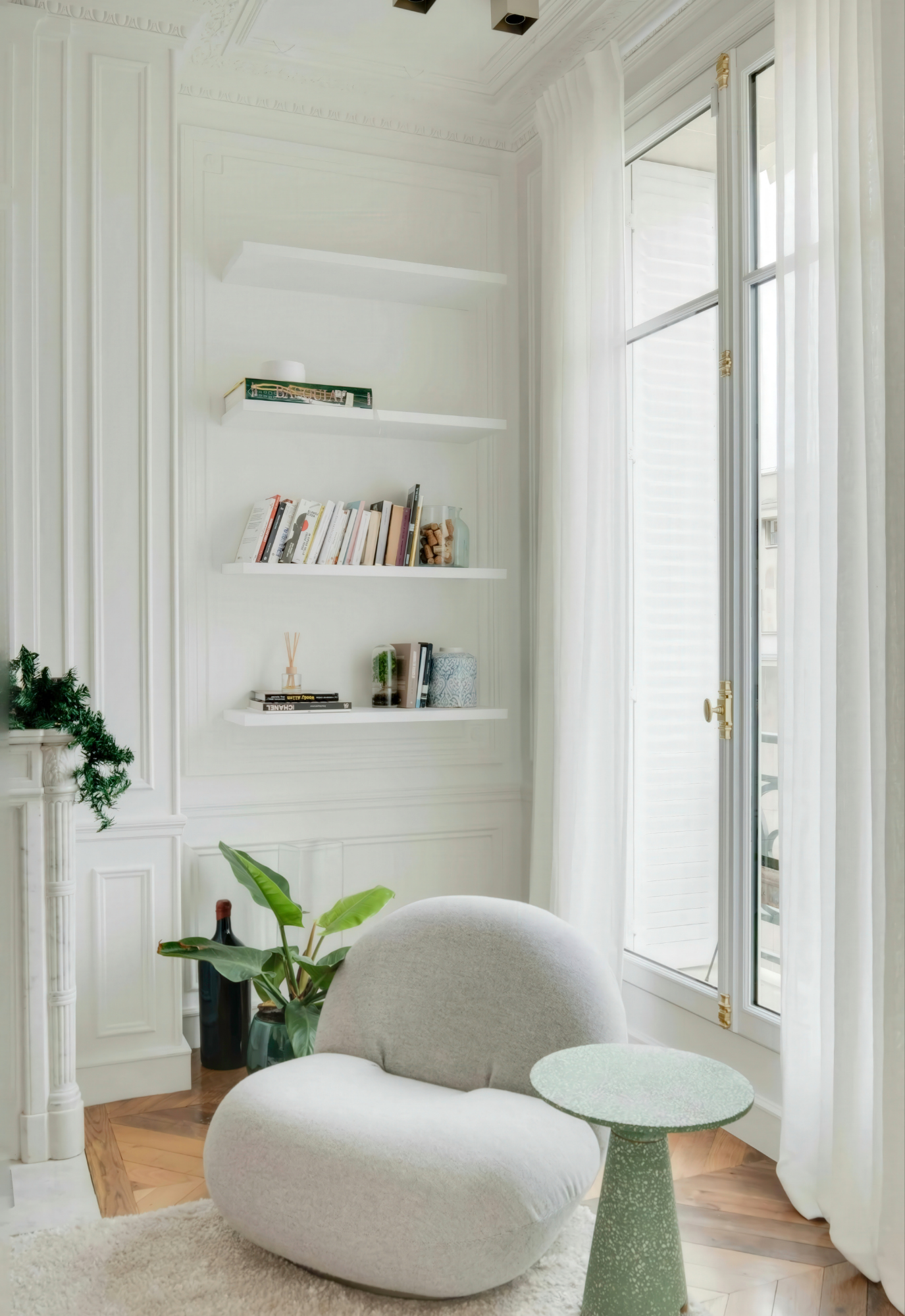 A cozy room with white walls, large window with sheer curtains, a beige rounded armchair, a green textured side table, indoor plants, and white built-in shelves with books and decor.