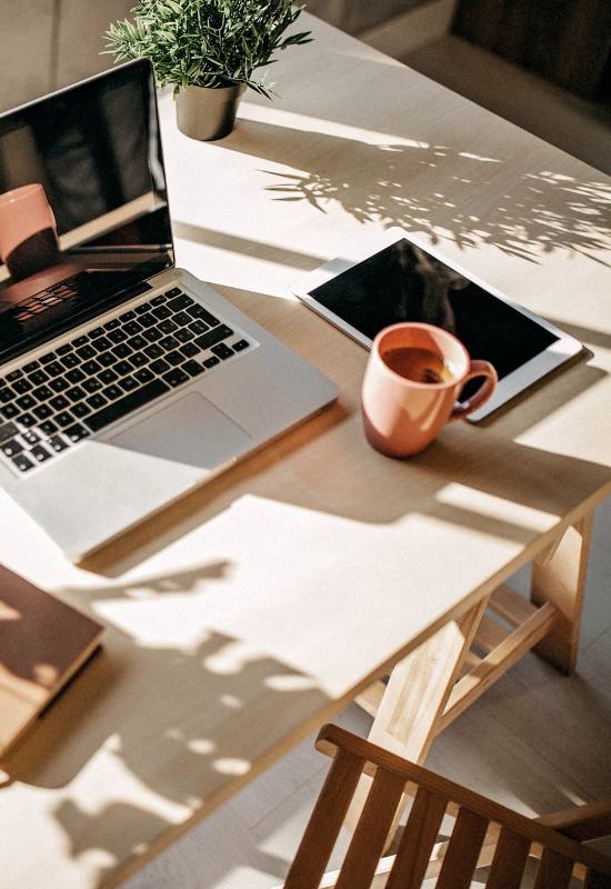 A wooden desk with an open laptop, a tablet, a pink coffee mug, and a potted plant, with sunlight casting shadows.