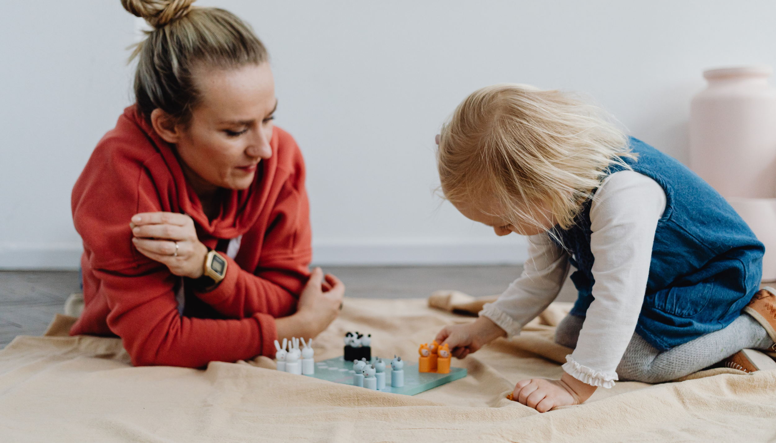 A woman and a young girl playing a game on the floor, both leaning in and focused on the game. The woman is wearing a red hoodie, and the girl is wearing a denim vest over a white long sleeve shirt. There's a beige blanket or sheet underneath them.