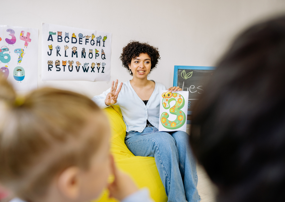 A teacher in a classroom showing a large decorative number three to children.