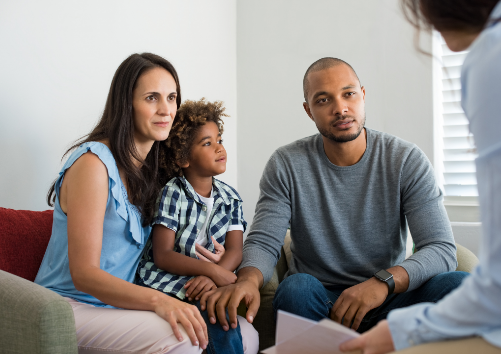 A family of three, including a woman, a young girl, and a man, sitting on a couch during a consultation with a professional in a brightly lit room.