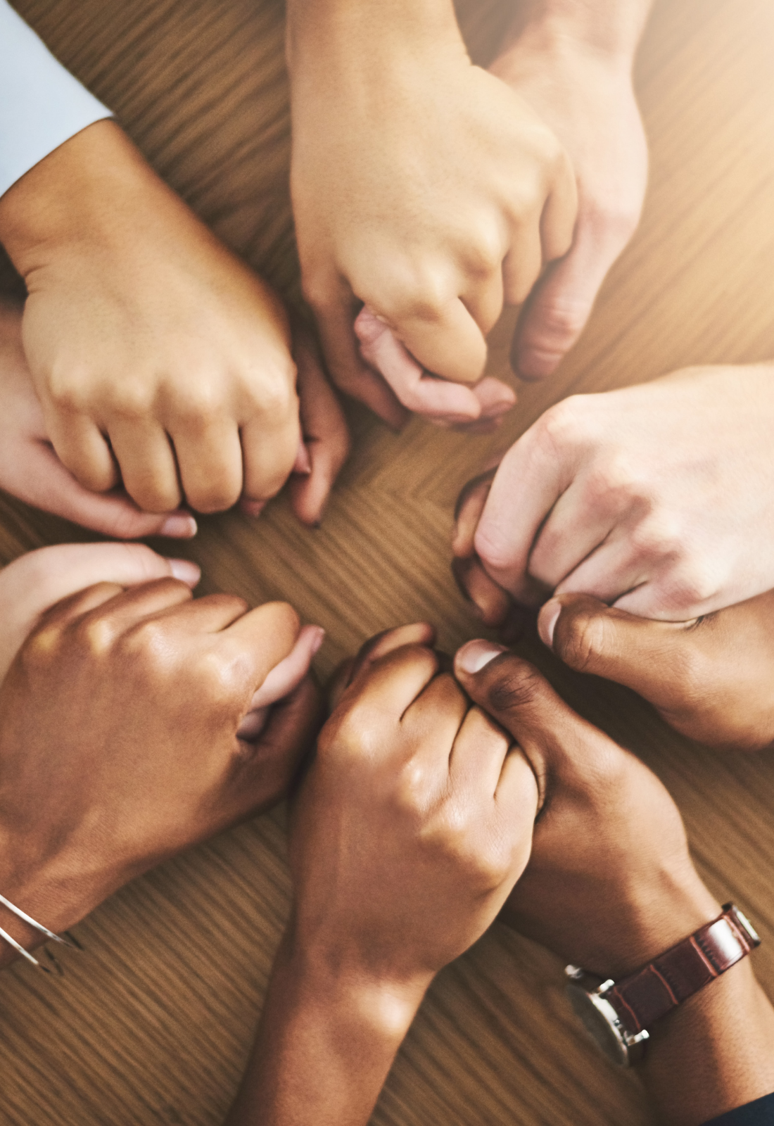 Multiple hands of diverse skin tones clasped together in a circle on a wooden surface.