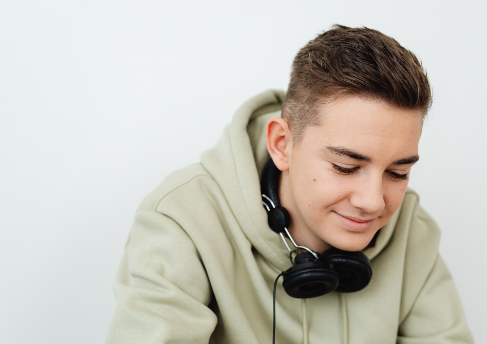 Young man with short hair wearing a beige hoodie and black headphones around his neck, smiling with his eyes closed, against a plain white background.
