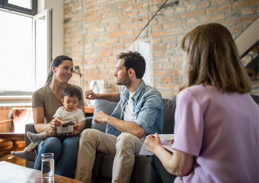 A family therapy session with a woman, a man, a young boy, and a counselor in a living room with a brick wall and large window.