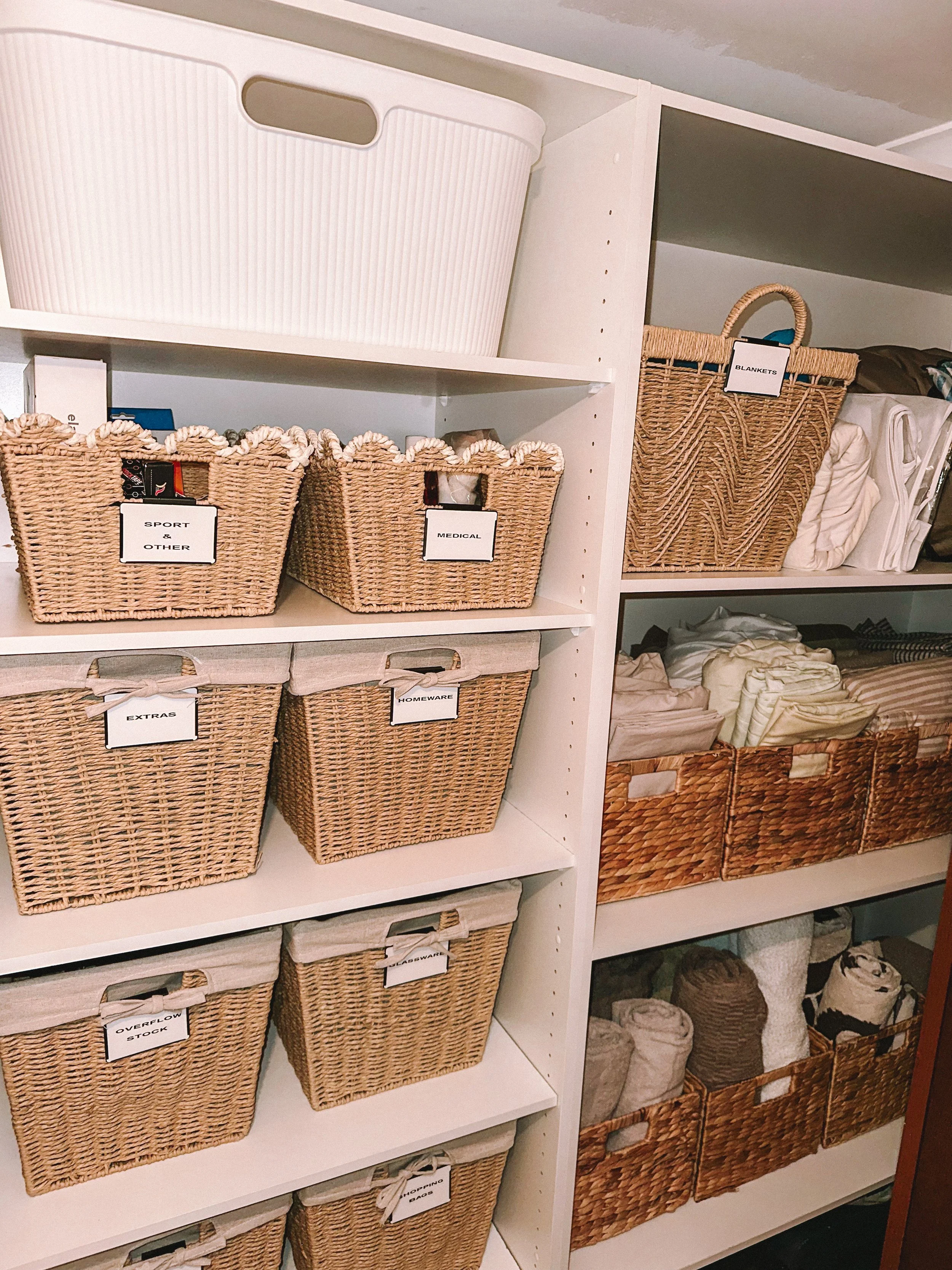White shelving unit with woven baskets labeled for storing various household items such as 'Sport & Other,' 'Medical,' 'Extras,' 'Glassware,' 'Overflow Stock,' and 'Shopping Bags.'