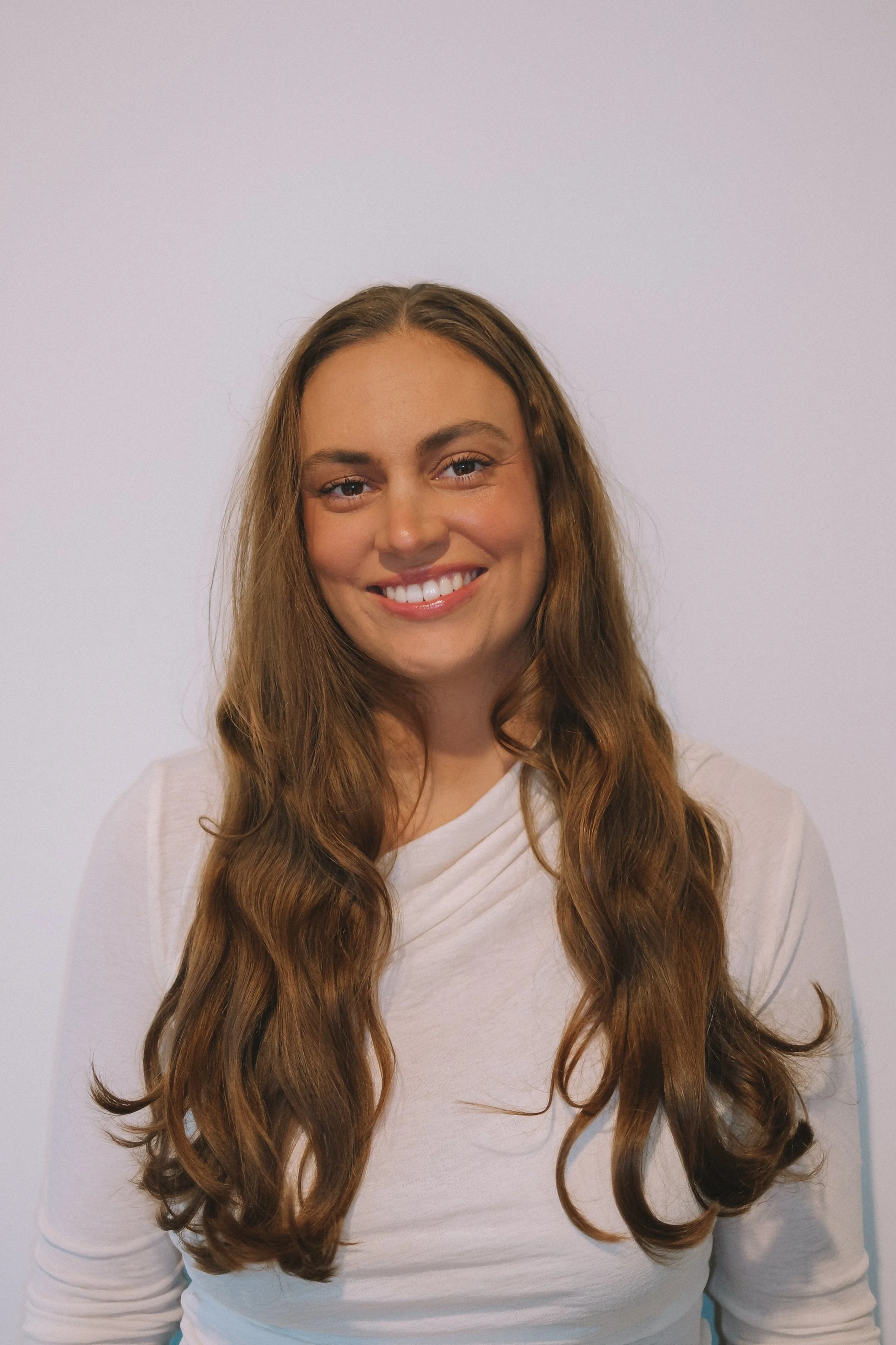 Close-up of a smiling woman with long wavy brown hair wearing a white top, standing against a plain light gray wall.