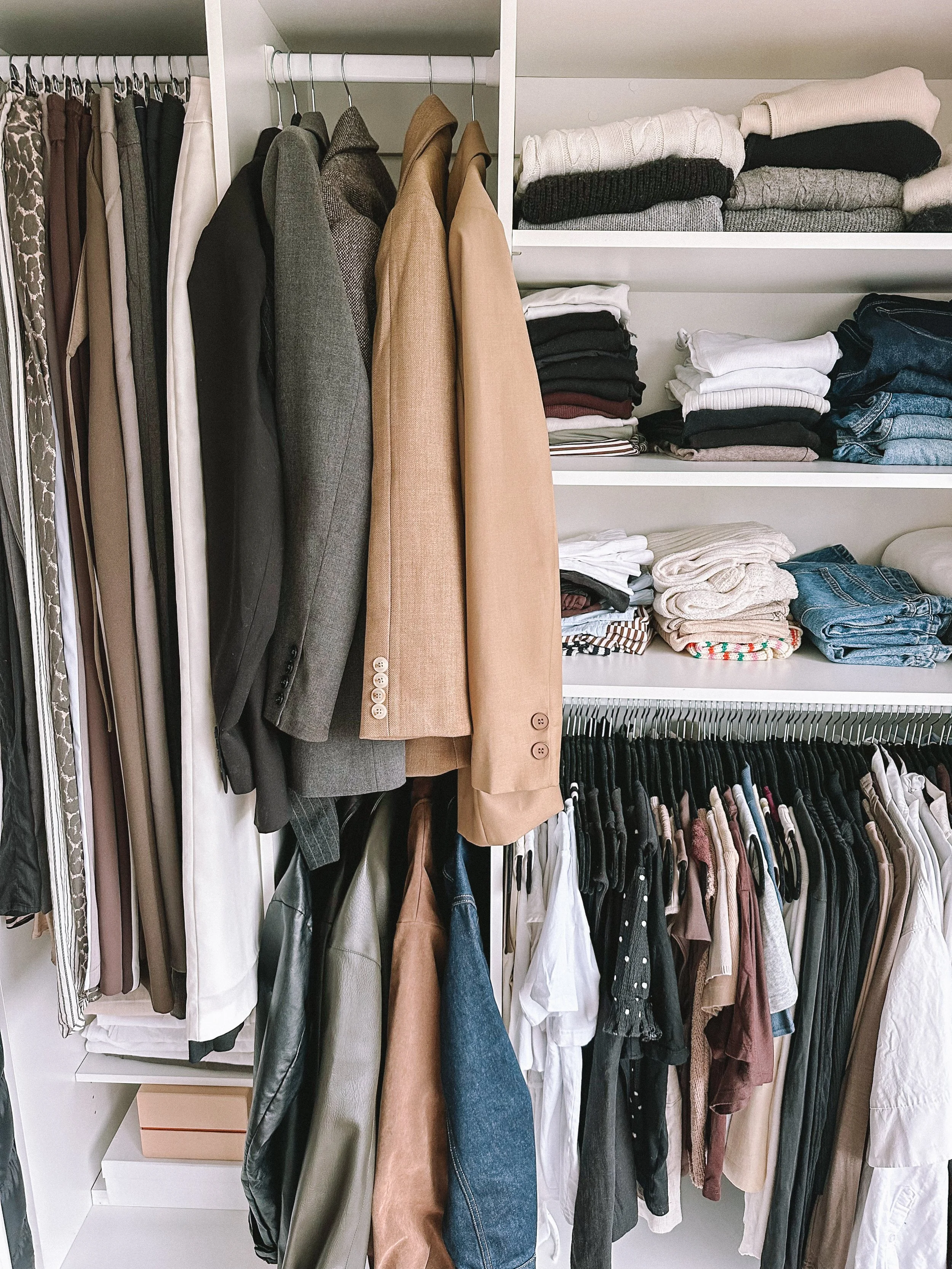 Clothing wardrobe with hanging blazers, shirts, and pants, and folded sweaters and jeans on shelves.