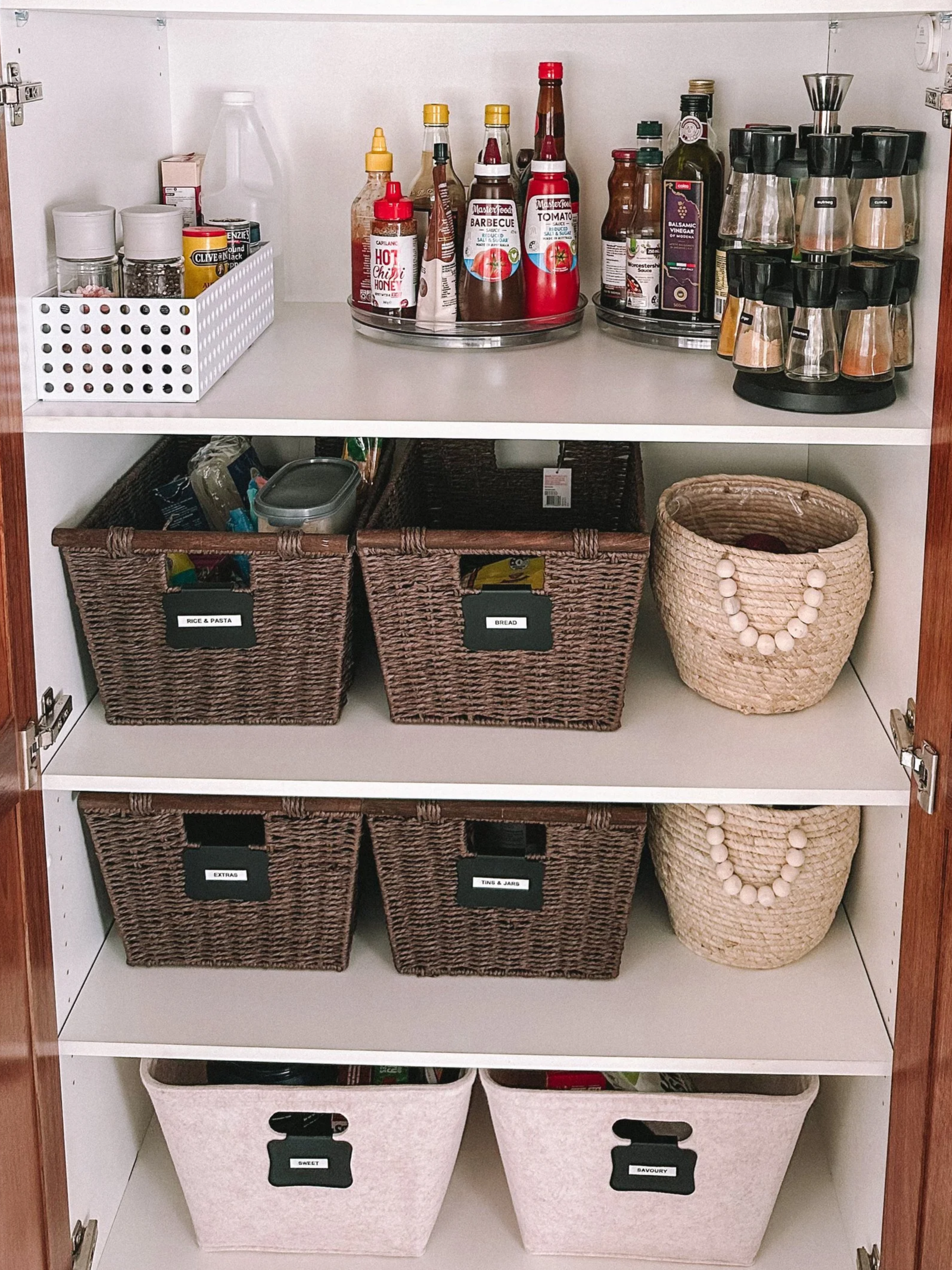 Inside a kitchen cabinet, there are various storage baskets and containers. The top shelf holds bottles of sauces, spices, and condiments. The middle shelf has three baskets labeled 'Rice & Pasta,' 'Bread,' and 'Extras,' along with a large decorative basket with a bead necklace. The bottom shelf contains two white storage bins labeled 'Sweet' and 'Savory.'