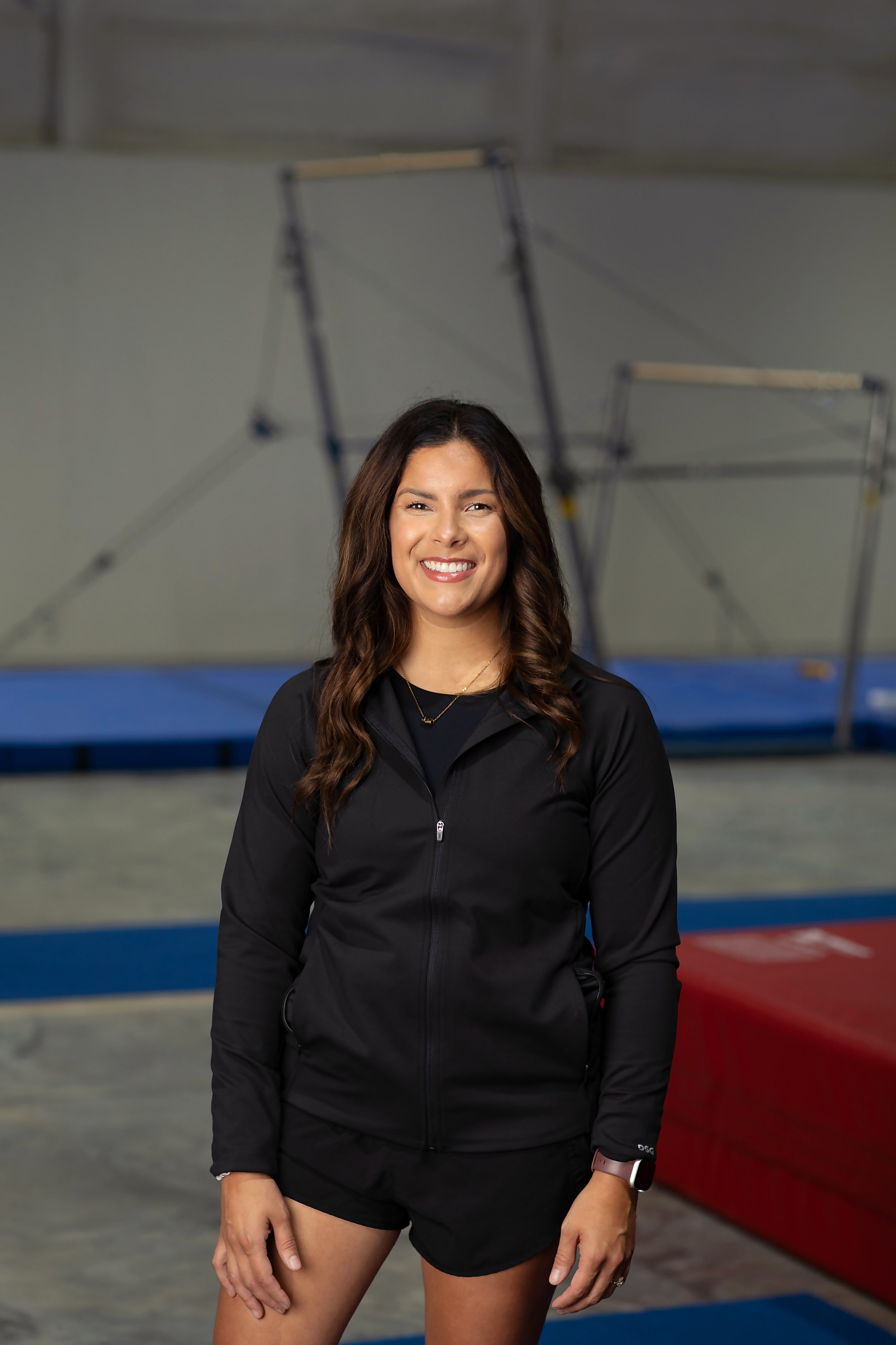 A woman smiling standing in an indoor gym with mats and equipment in the background.