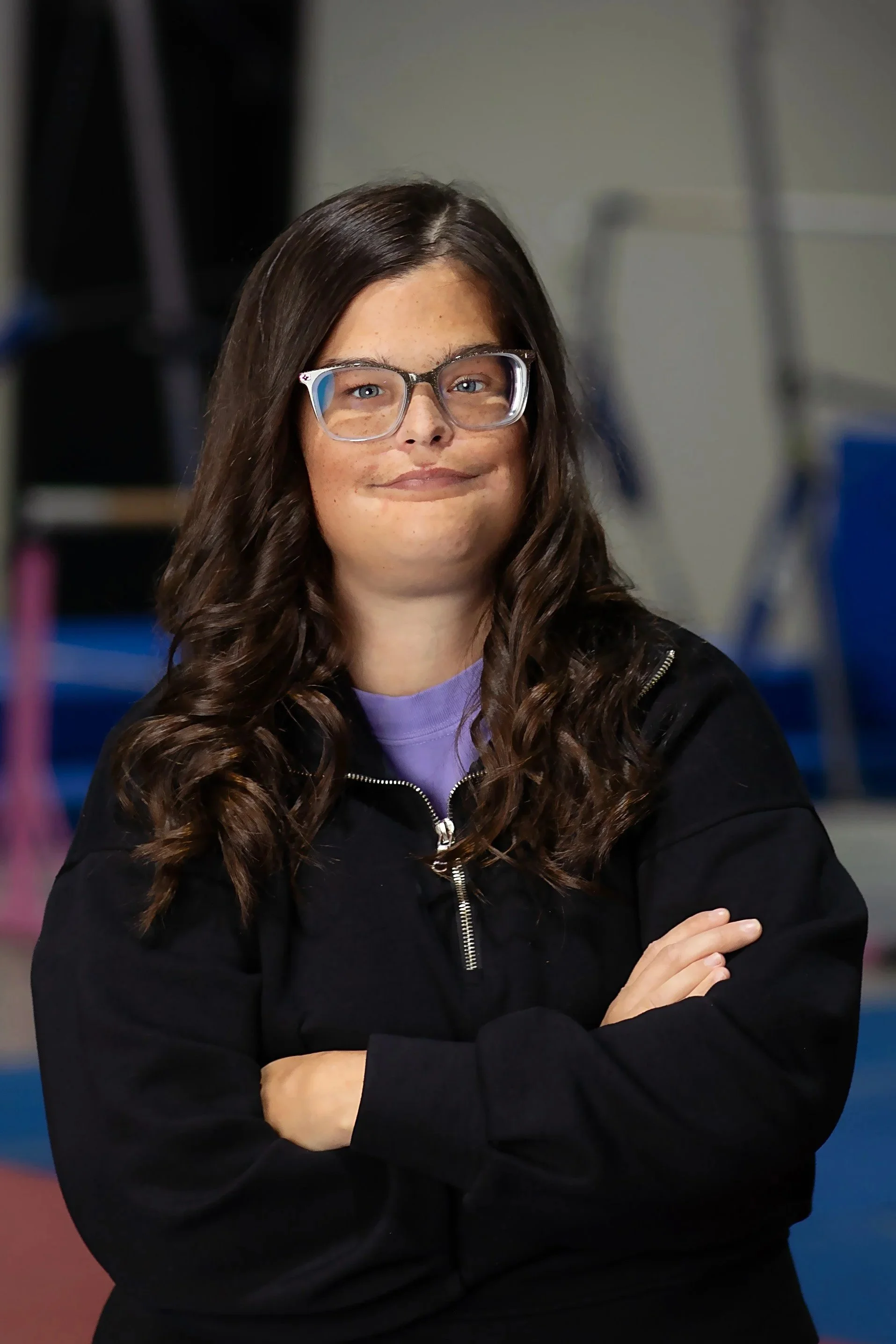 A woman with long, curly brown hair, wearing glasses, a purple shirt, and a black zip-up jacket, standing with arms crossed in an indoor setting.
