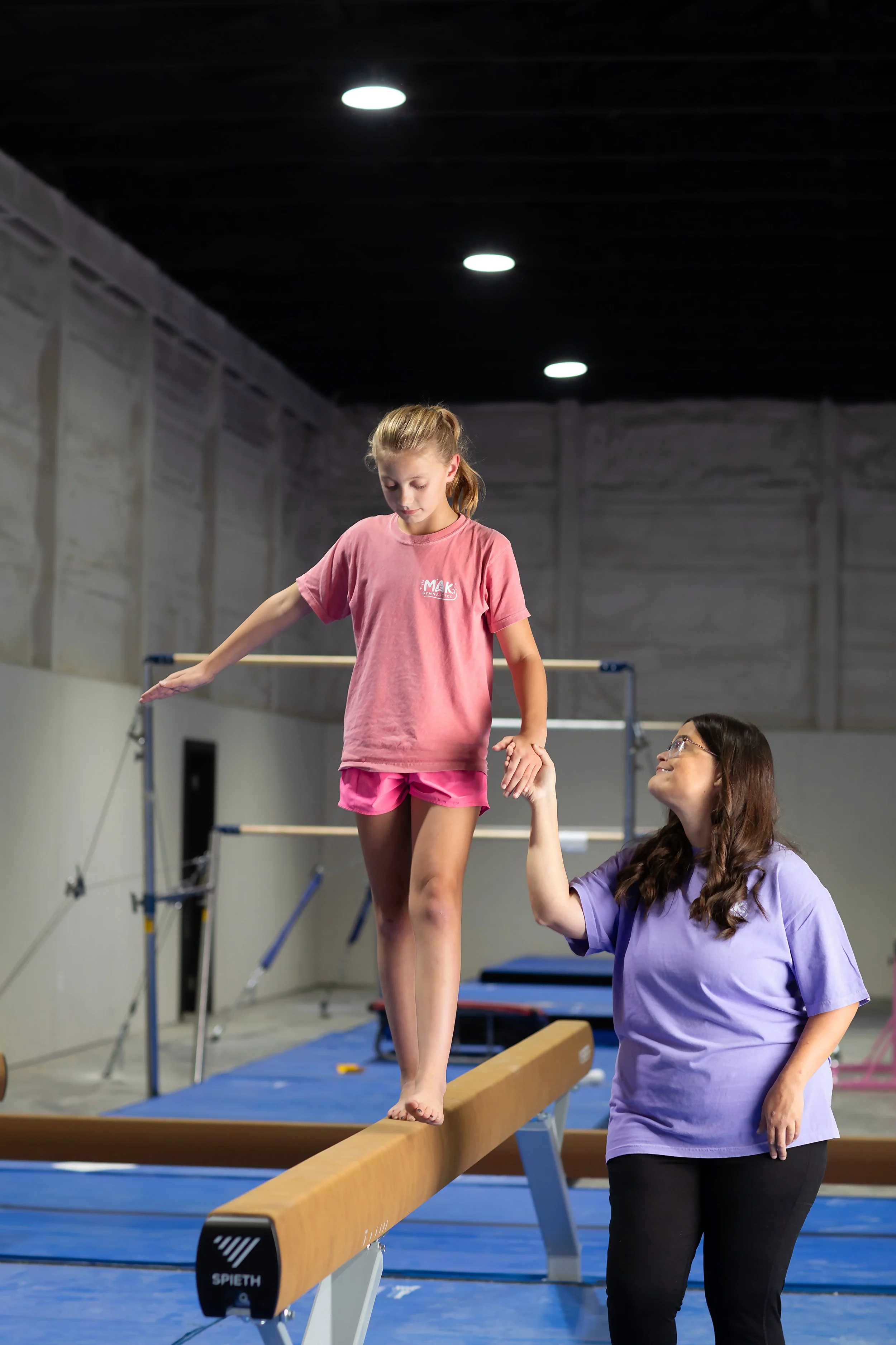 A young girl dressed in pink is balancing on a wooden beam with assistance from a woman in a purple shirt. The setting is an indoor gymnastics training facility with mats and equipment.