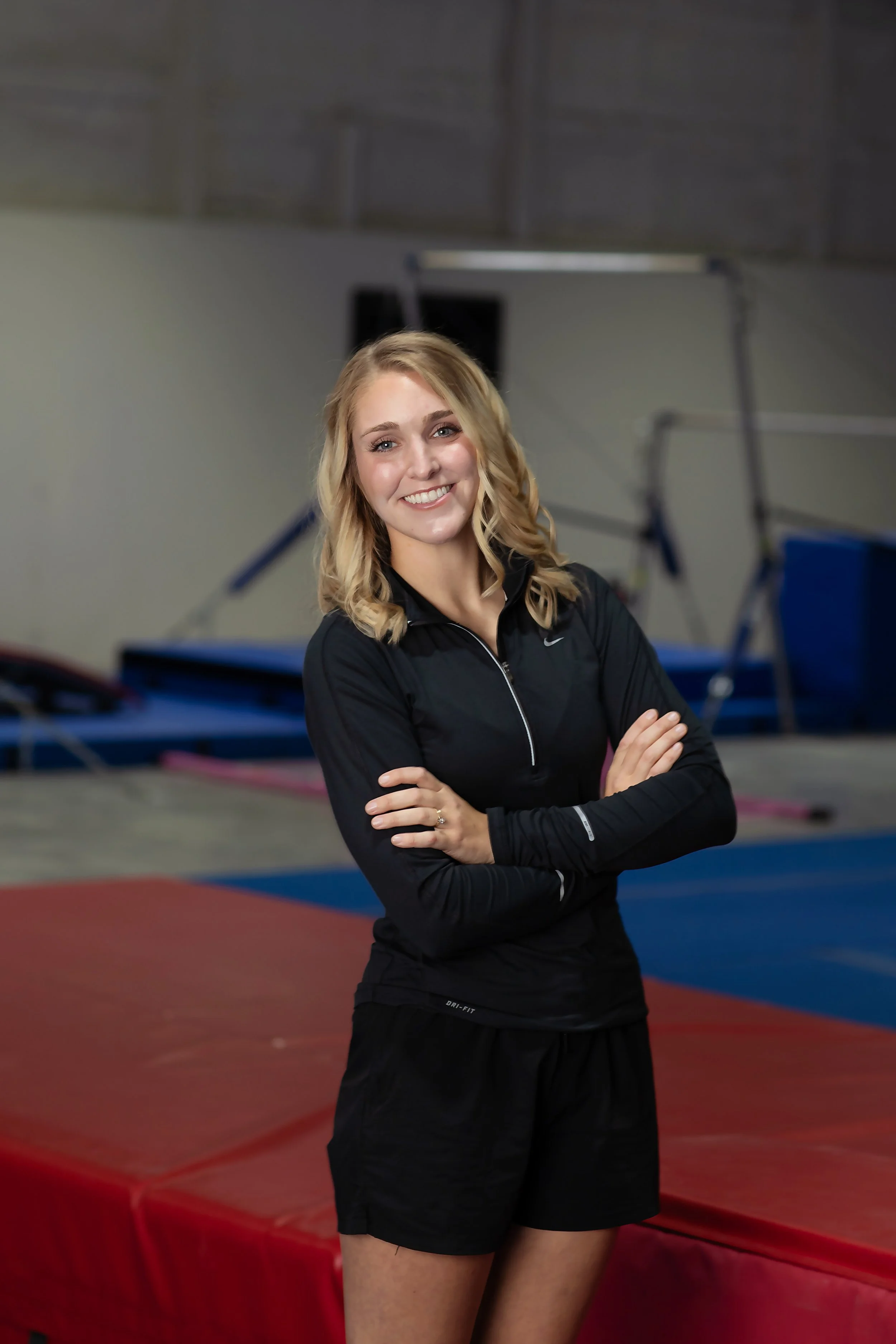 A young woman with blonde hair and a black athletic shirt standing with her arms crossed in an indoor gym, with gymnastics equipment in the background.