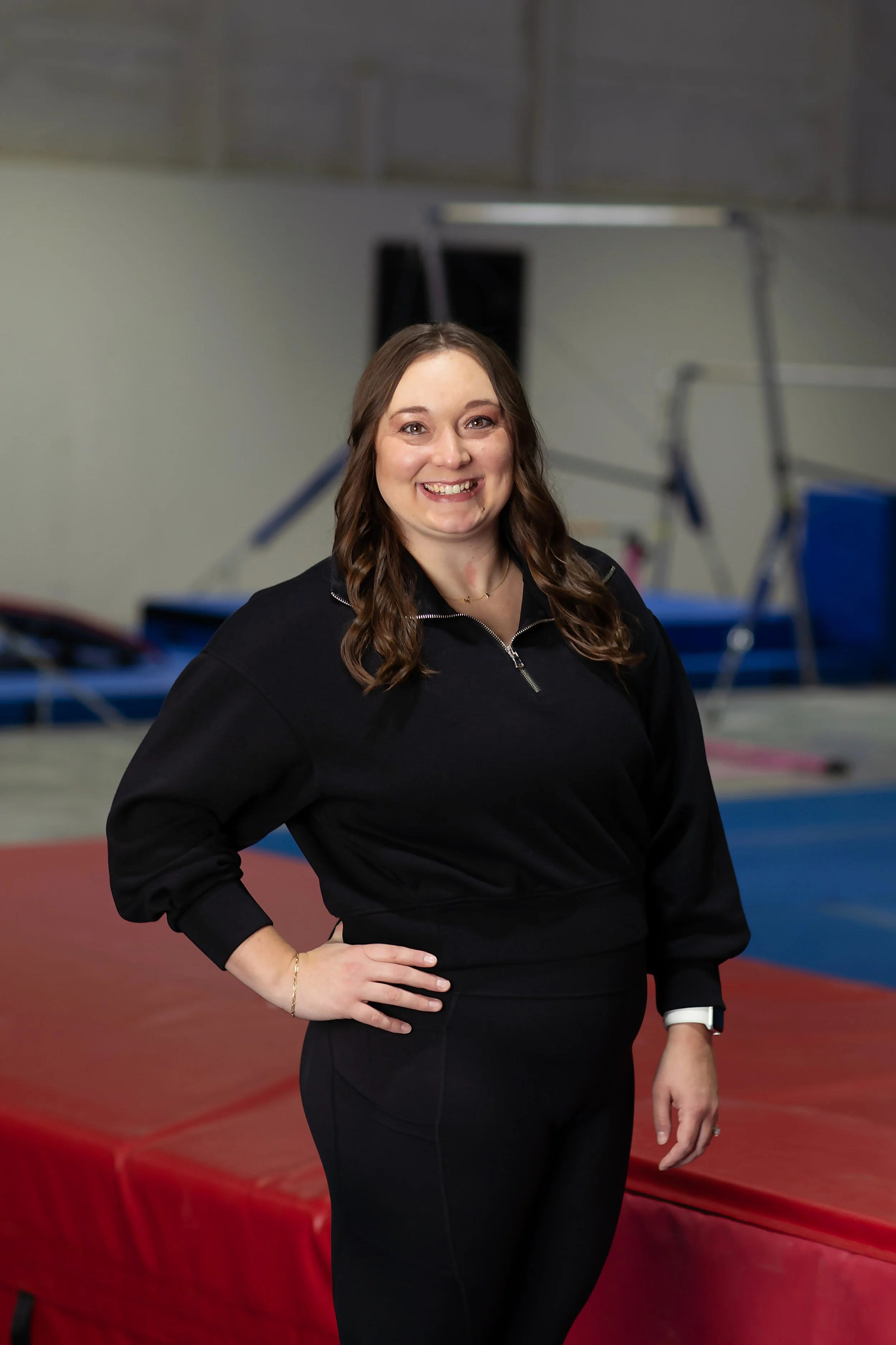 A woman with brown curly hair smiling, wearing a black zip-up top, standing with her right hand on her hip in what appears to be an indoor gym or sports facility with gym equipment and mats in the background.