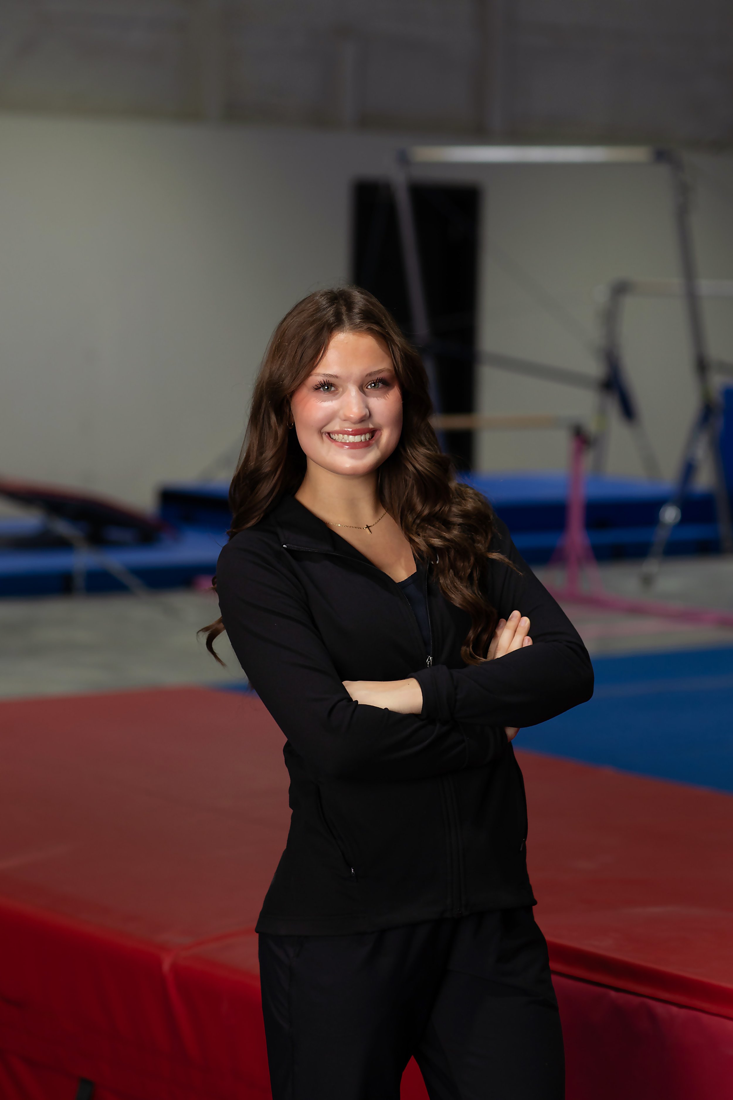 Young woman with long brown hair smiling and standing with arms crossed in an indoor gymnastics facility, with mats and gymnastic equipment in the background.