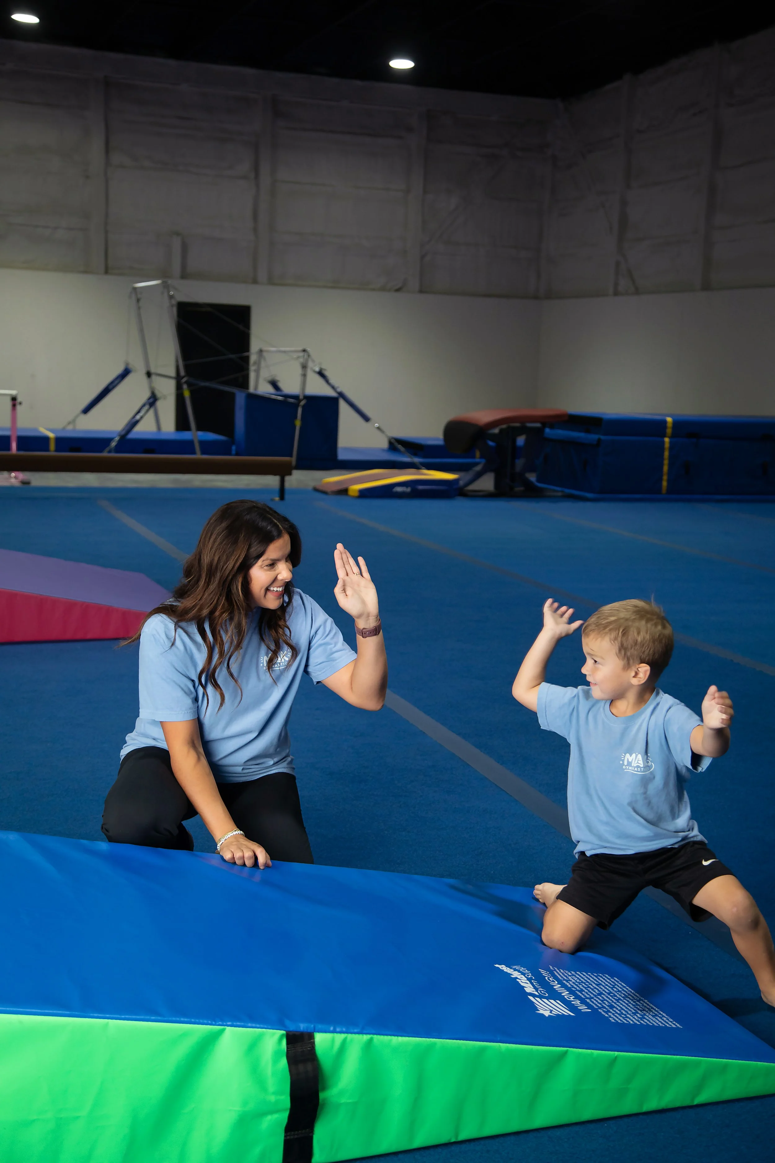 A young boy and a woman sitting on the floor of a gymnastics facility, smiling and giving high-fives. The background shows gymnastic mats and equipment.