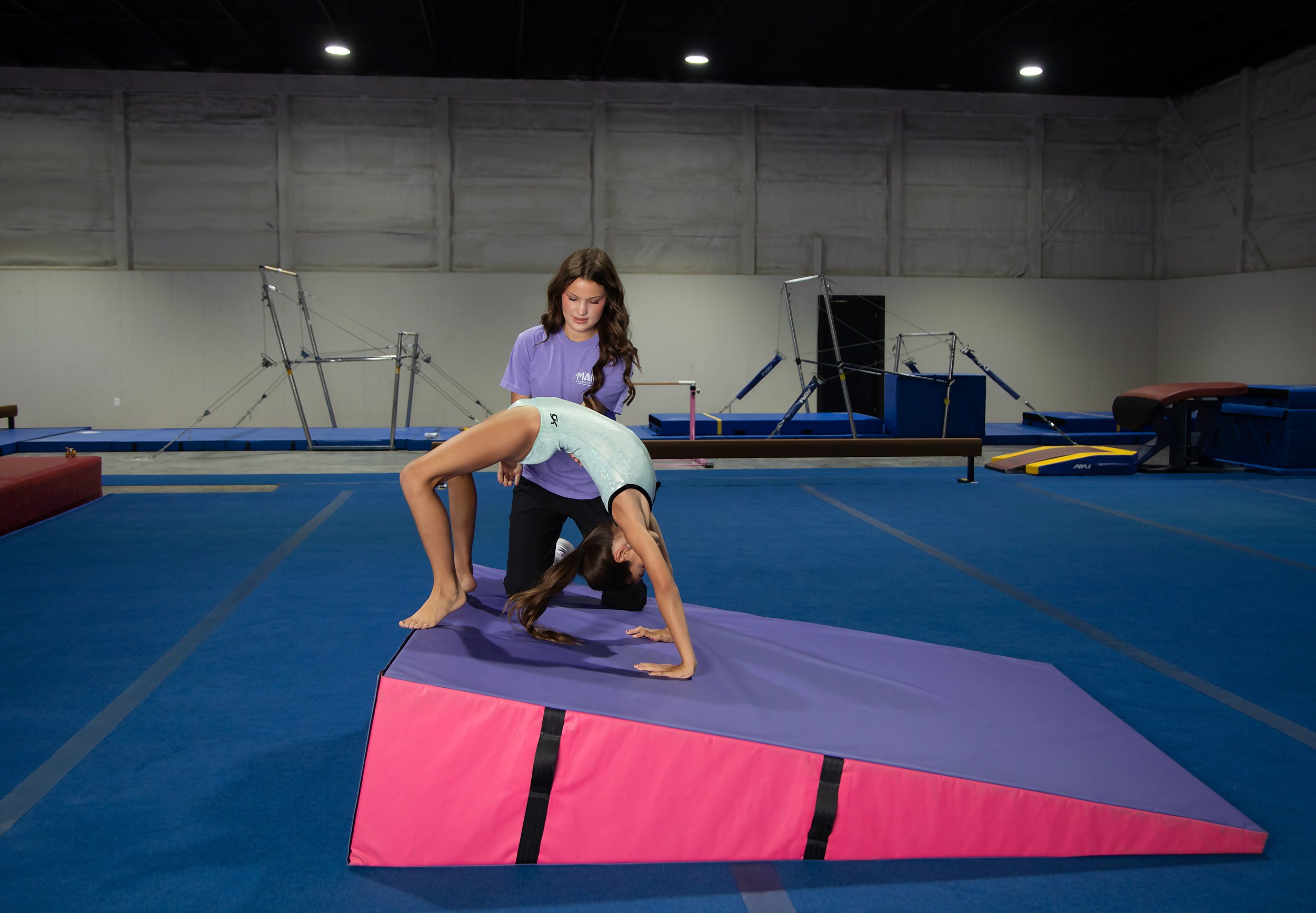 A young woman in a purple t-shirt assisting another woman in a black outfit with her back arched on a pink and purple gymnastics mat inside a gymnasium. There are balance beams and gymnastics equipment in the background.