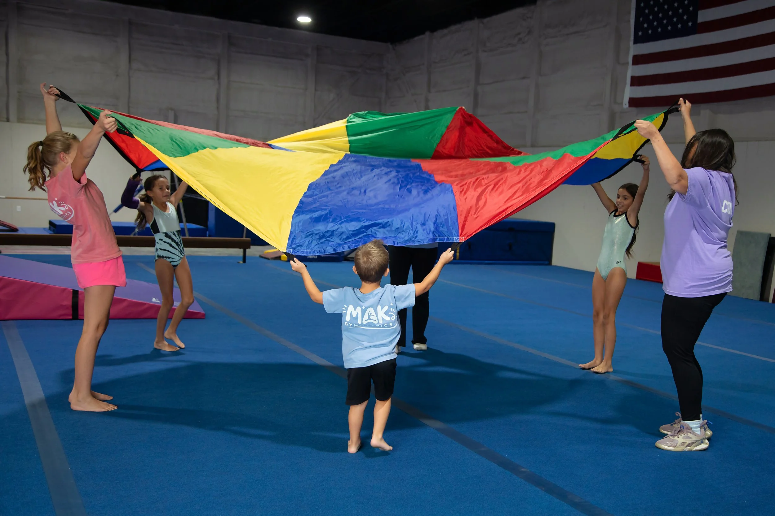 Children and an adult in a gymnasium holding a colorful parachute together, engaging in a group activity.