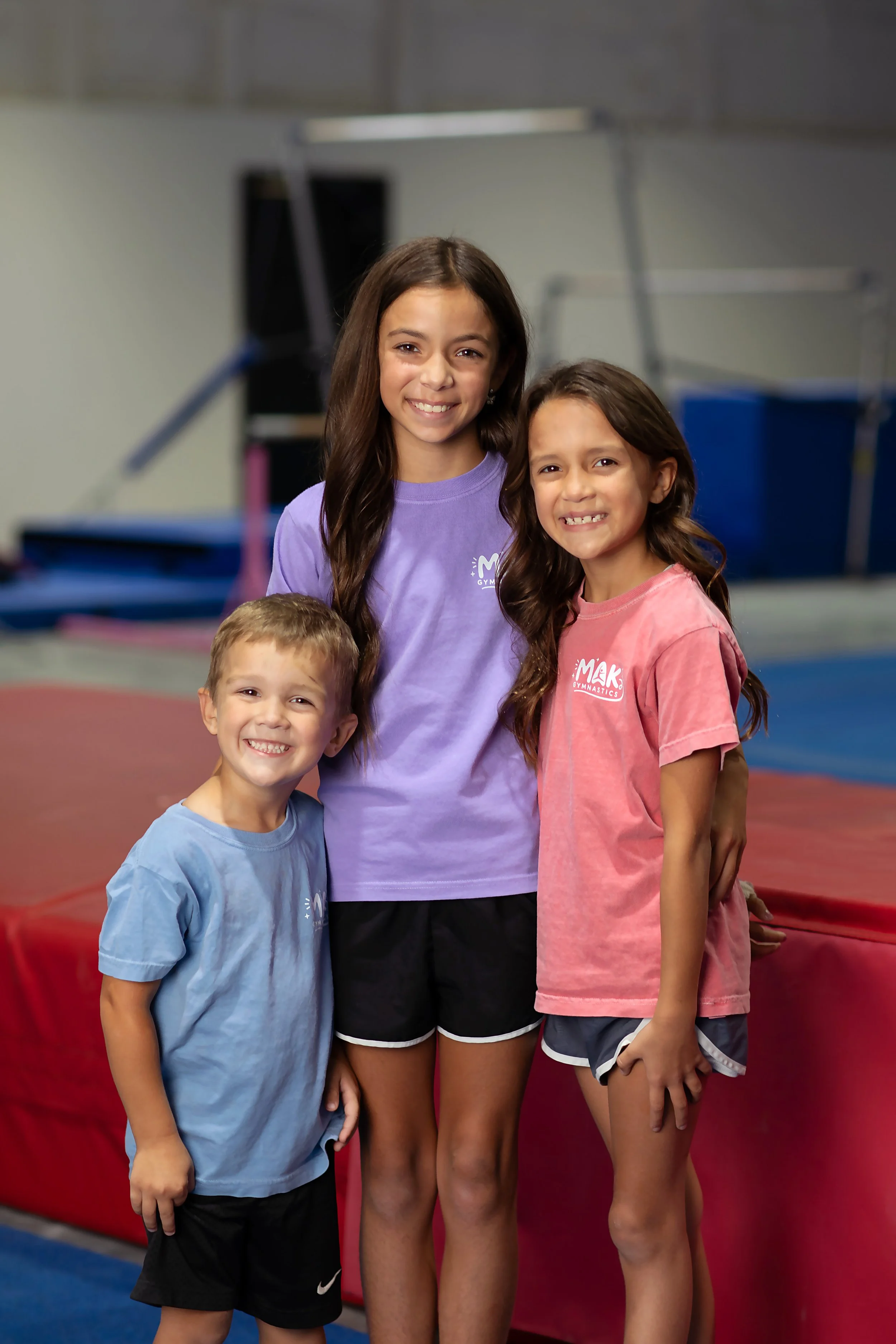 Four children in a gymnastics gym, smiling. Three girls and one boy, wearing colorful T-shirts with gym logos, standing in front of gymnastic equipment like a springboard and mats.