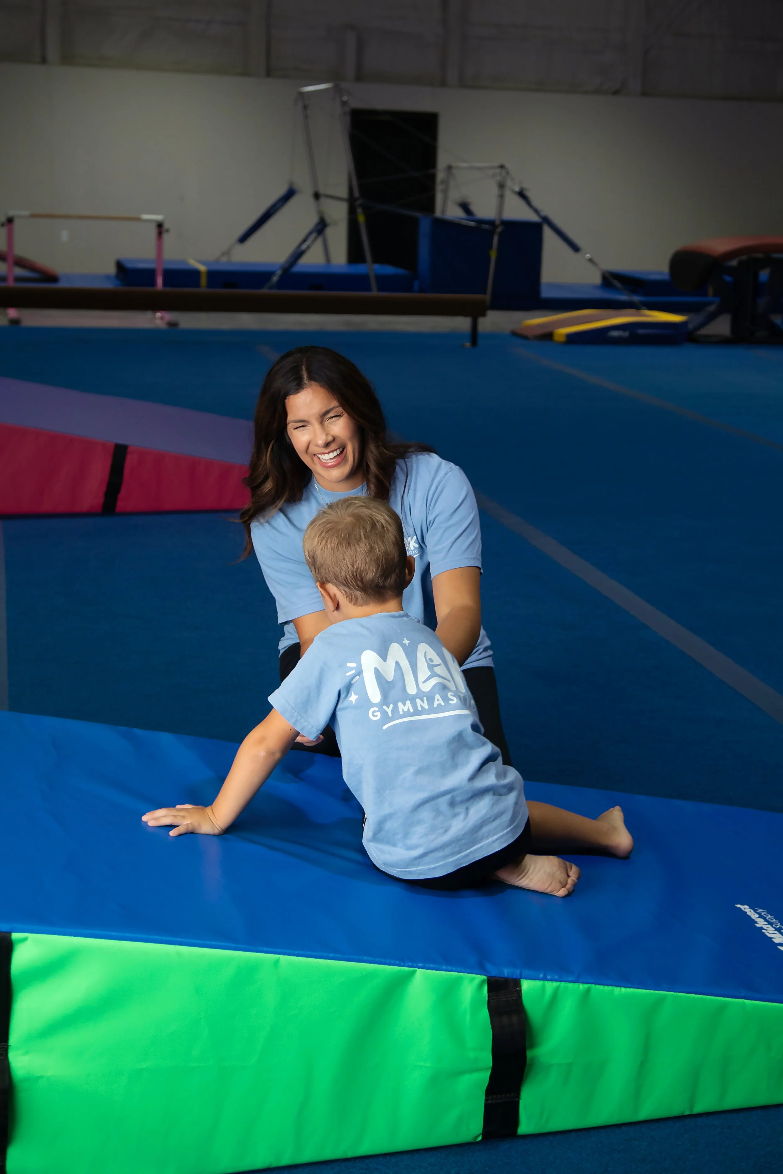 Young woman smiling and talking to young boy on a gymnastics mat in an indoor gymnasium.