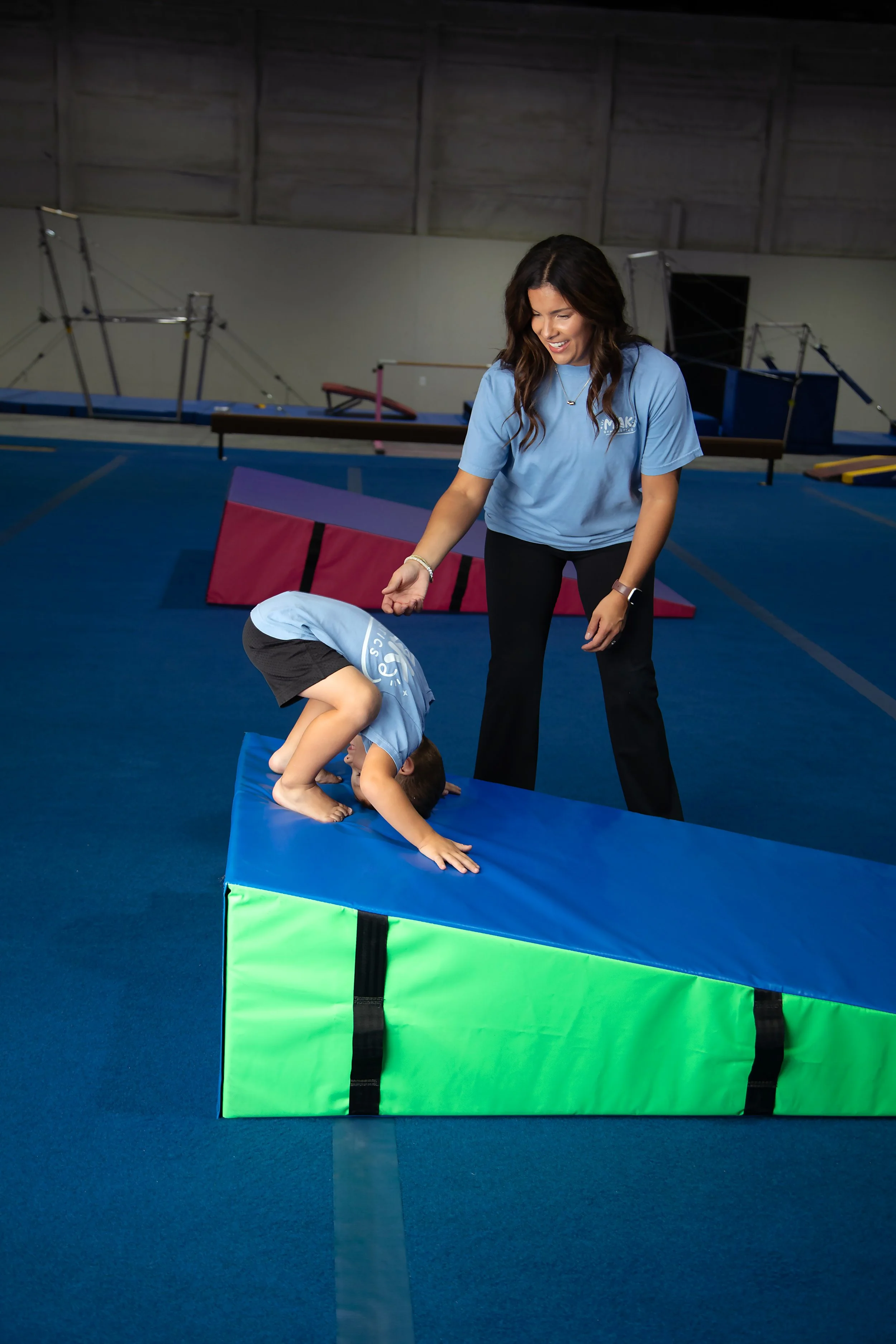 A young girl is bending over on a padded gym mat, with her hands and feet on the mat, and her head tucked under her arm. A woman, possibly a coach or instructor, stands smiling nearby, watching her. The setting is an indoor gymnastics gym with equipment in the background and a blue floor.
