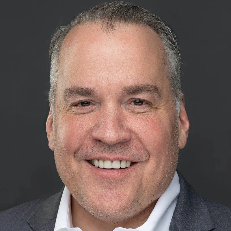 A close-up portrait of a smiling man with short gray hair, dressed in a dark suit and white shirt, against a dark background.