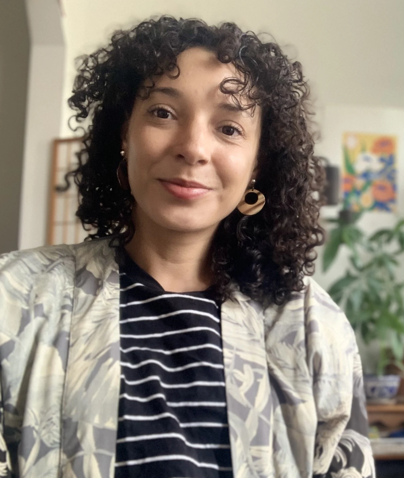 A woman with dark, curly hair, wearing earrings and a patterned blazer over a black and white striped shirt, taking a selfie in a room with a colorful abstract painting on the wall and a plant in the background.