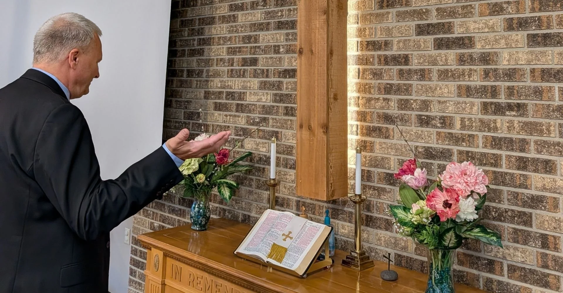 A man dressed in a black suit with a light blue shirt praying in front of an altar with flowers, candles, and an open Bible, inside a church with a brick wall background.