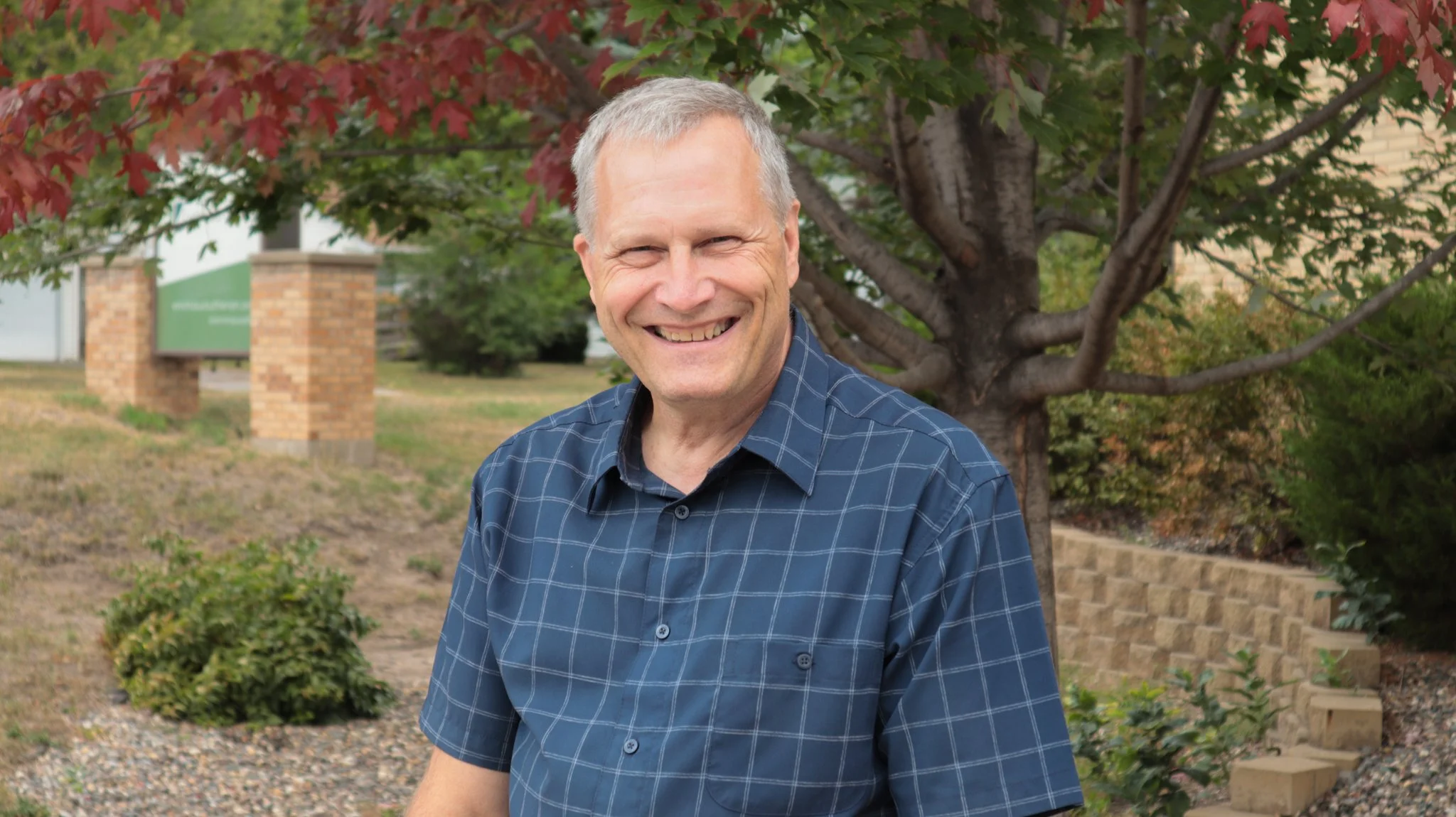 A smiling middle-aged man with gray hair, wearing a blue plaid shirt, standing outdoors near a tree with red and green leaves.