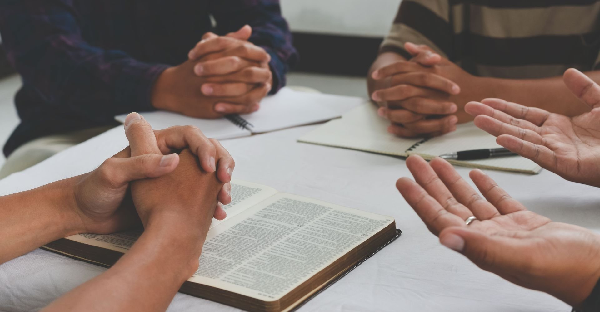 Group of people gathered around a table in prayer, with hands clasped and an open Bible visible on the table.