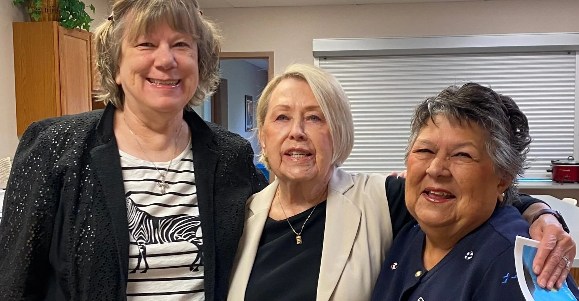 Three women standing together, smiling and embracing, indoors with a closed window and wooden cabinets in the background.