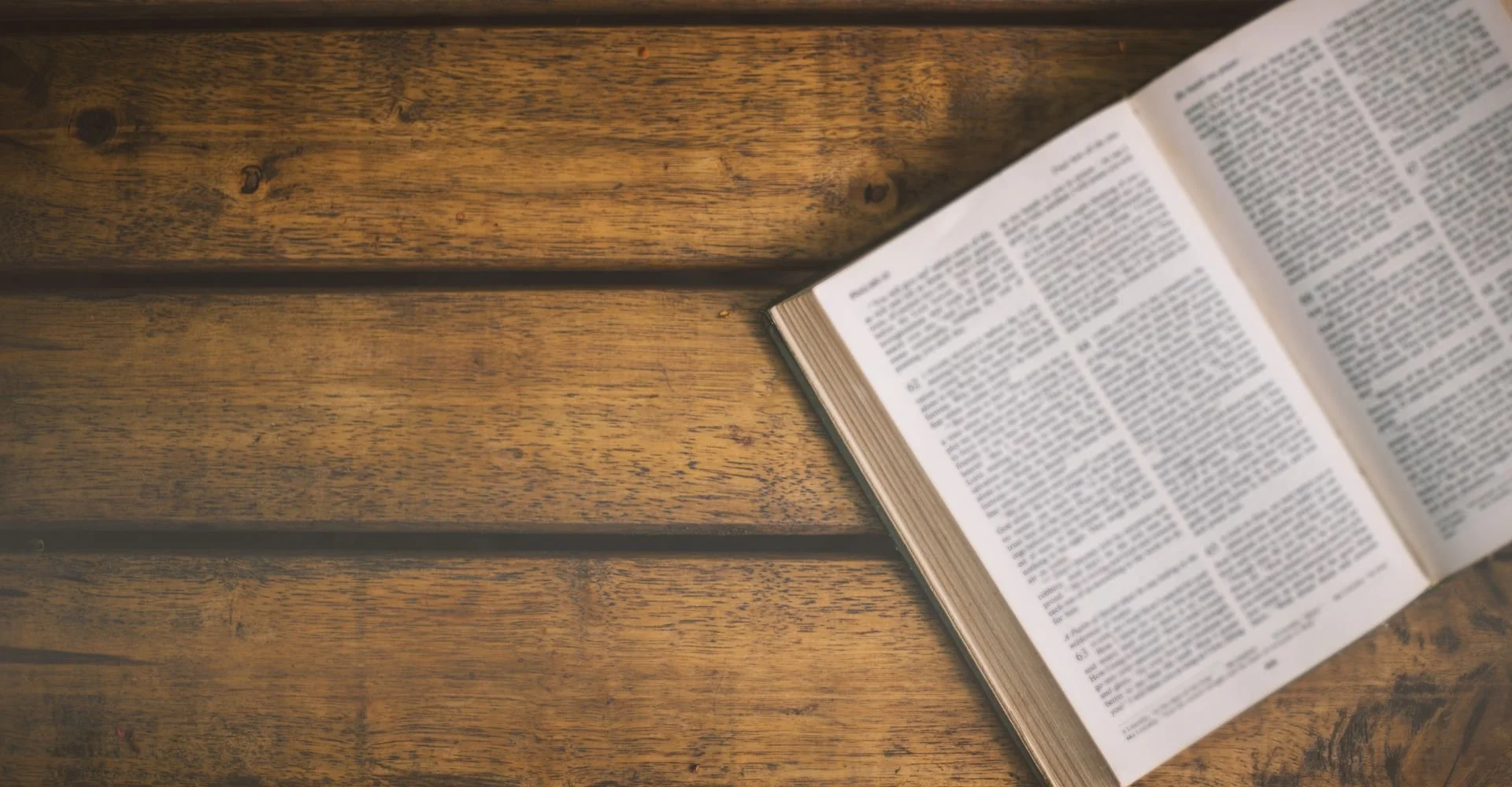 An open book with small text lies on a wooden surface with visible grains and knots.