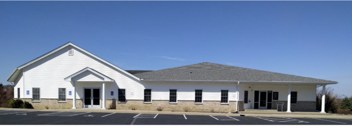A white building with brick base and multiple windows, featuring a gabled roof and a covered entrance with supporting columns, located in a parking lot under a clear blue sky.