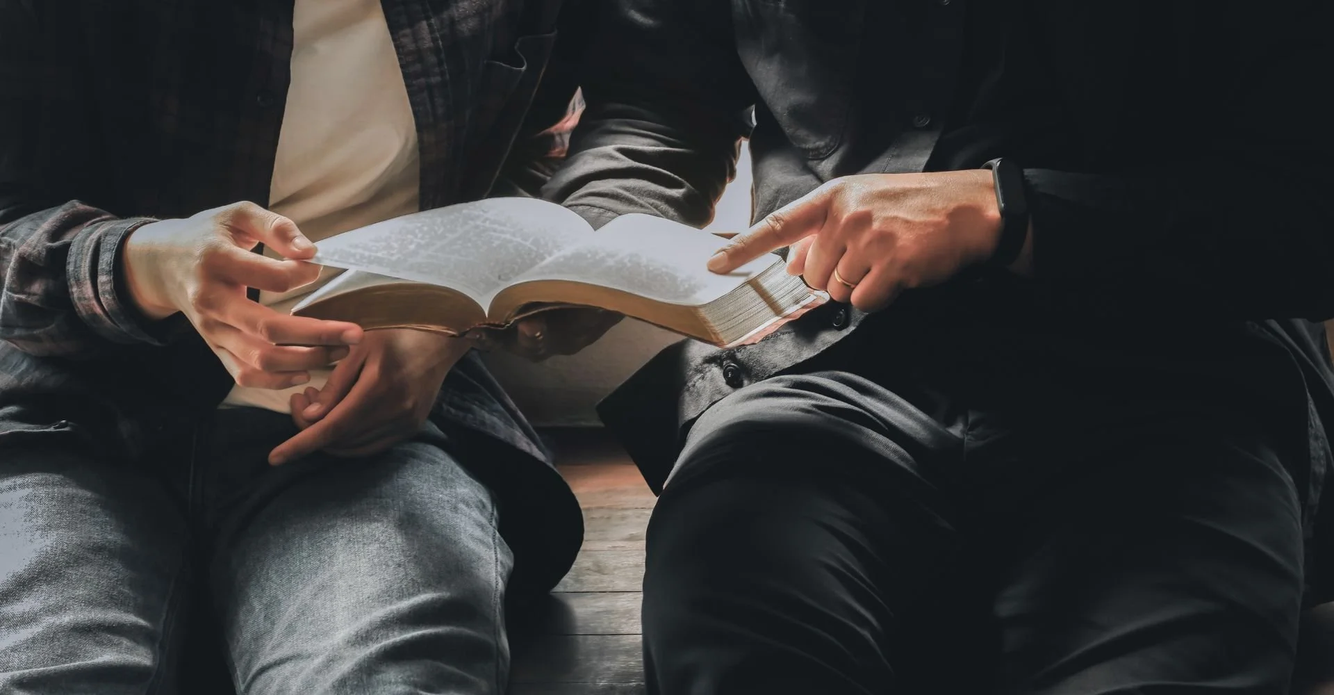 Two people sitting close together, reading a book on a dark wooden floor.