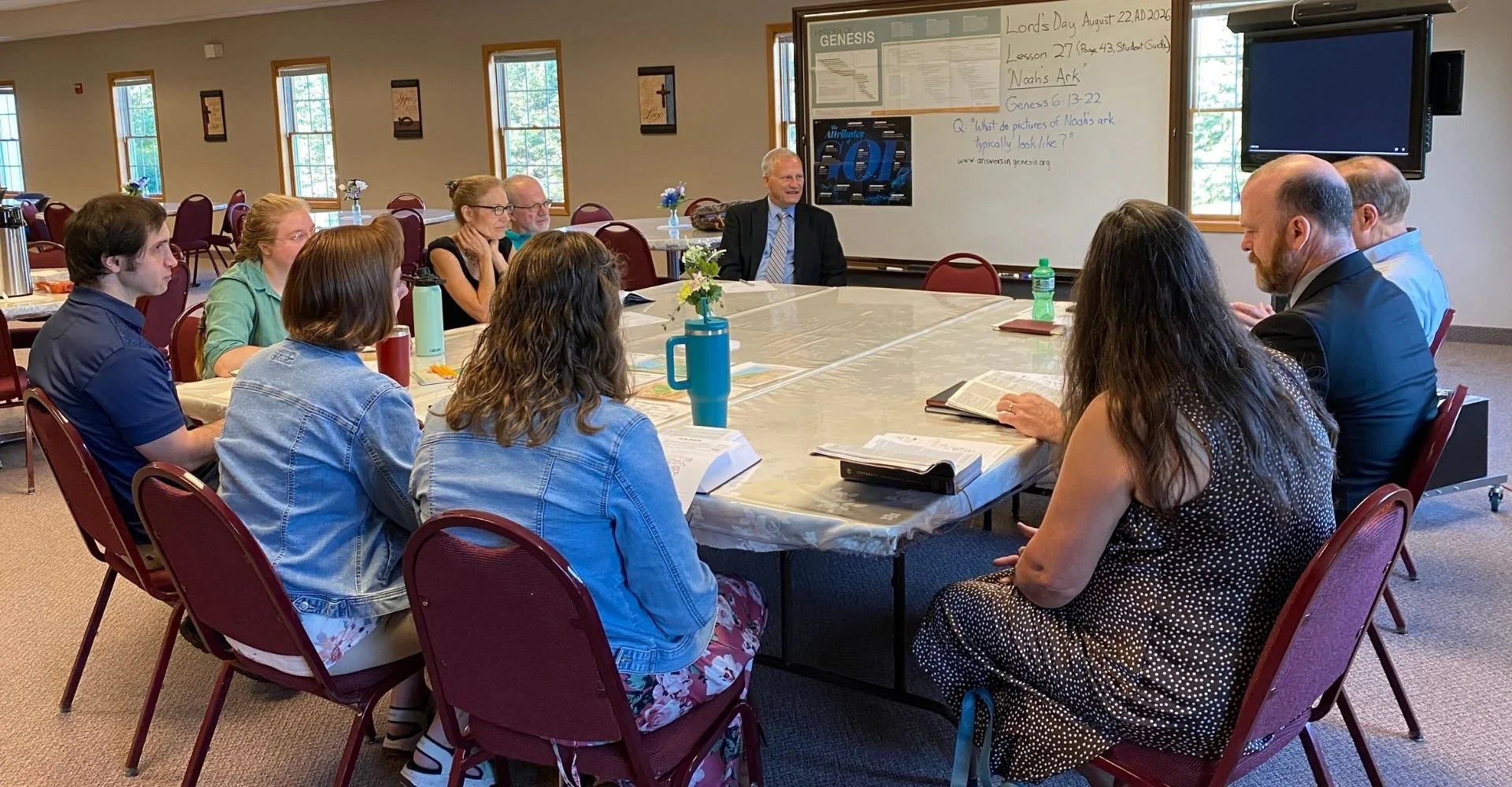 A group of people sitting around a large table in a conference room, participating in a discussion or class session. There are 10 individuals, some reading from open books, with a whiteboard, windows, and framed religious posters on the wall in the background.