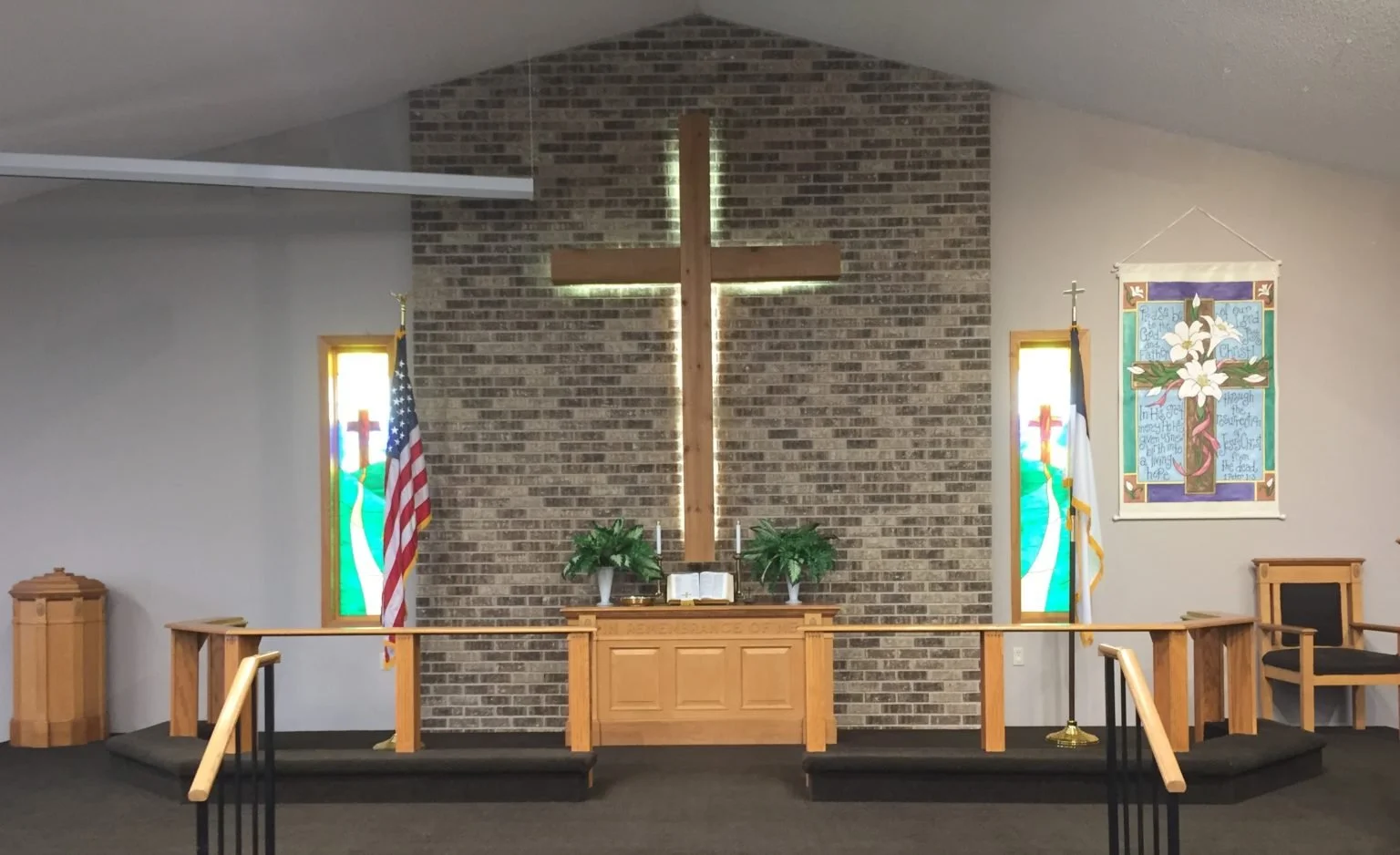 Interior of a church sanctuary with a large wooden cross on a brick wall behind the altar, flanked by two stained glass windows, two flags, and a wall hanging with lilies and biblical text. The altar has two potted plants and an open Bible, with a carpeted platform and wooden railing.