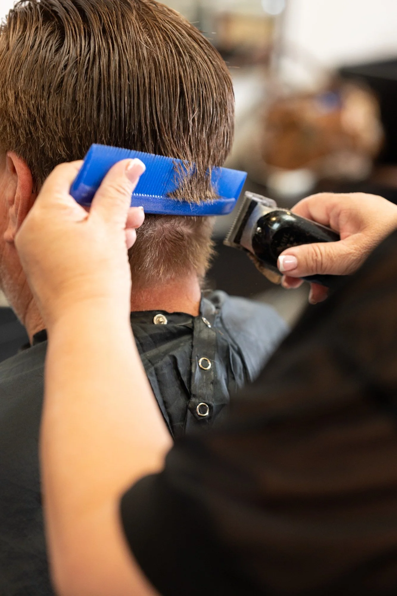 A person getting a haircut, with hair clippers trimming the back of their brown hair. The barber is holding a blue comb and using their hand to guide the hair. The individual is wearing a black cape with metallic buttons.