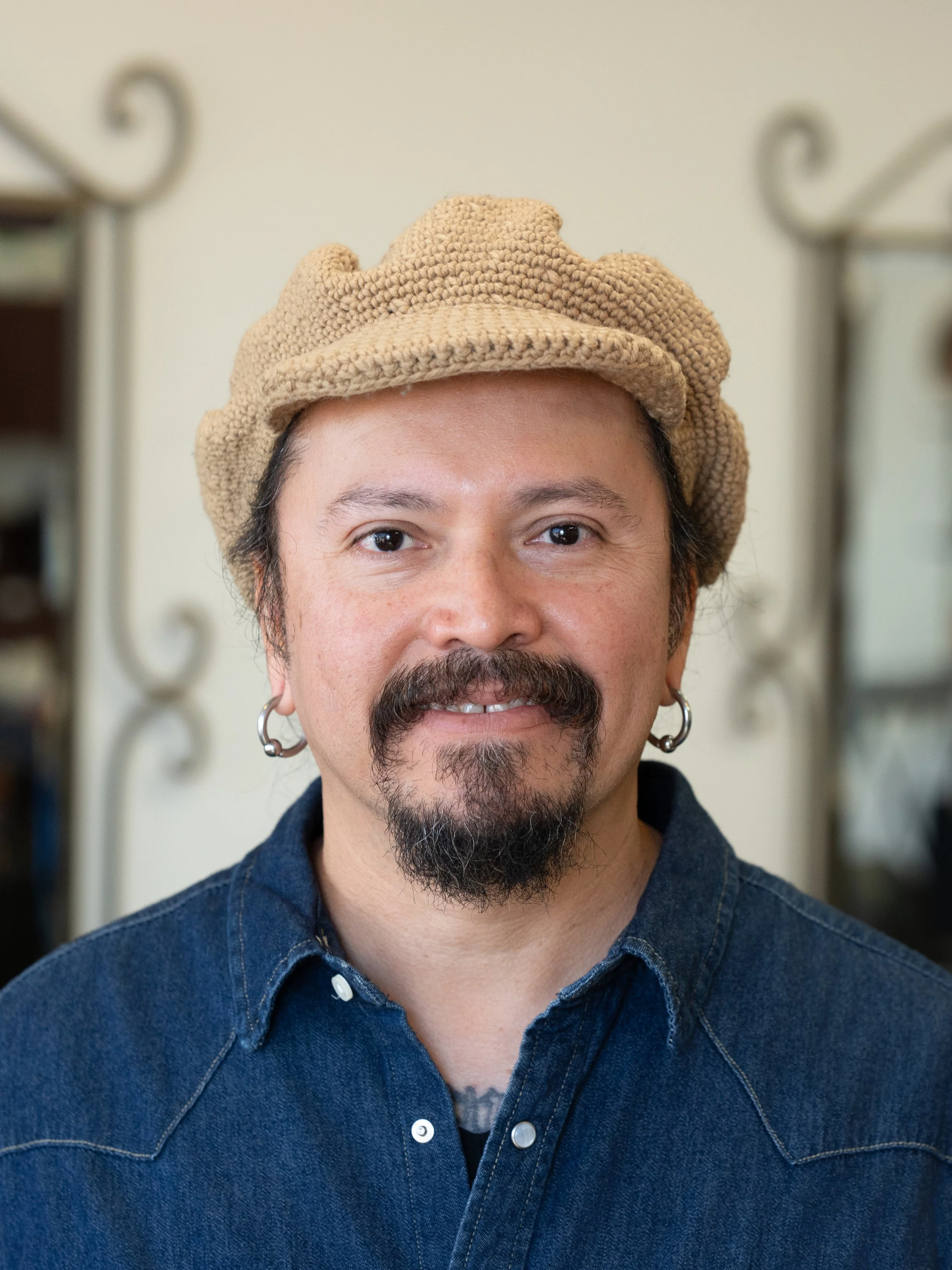 Close-up of a man wearing a tan knit hat and a dark blue denim shirt, with earrings and facial hair, smiling softly in front of a blurred background.