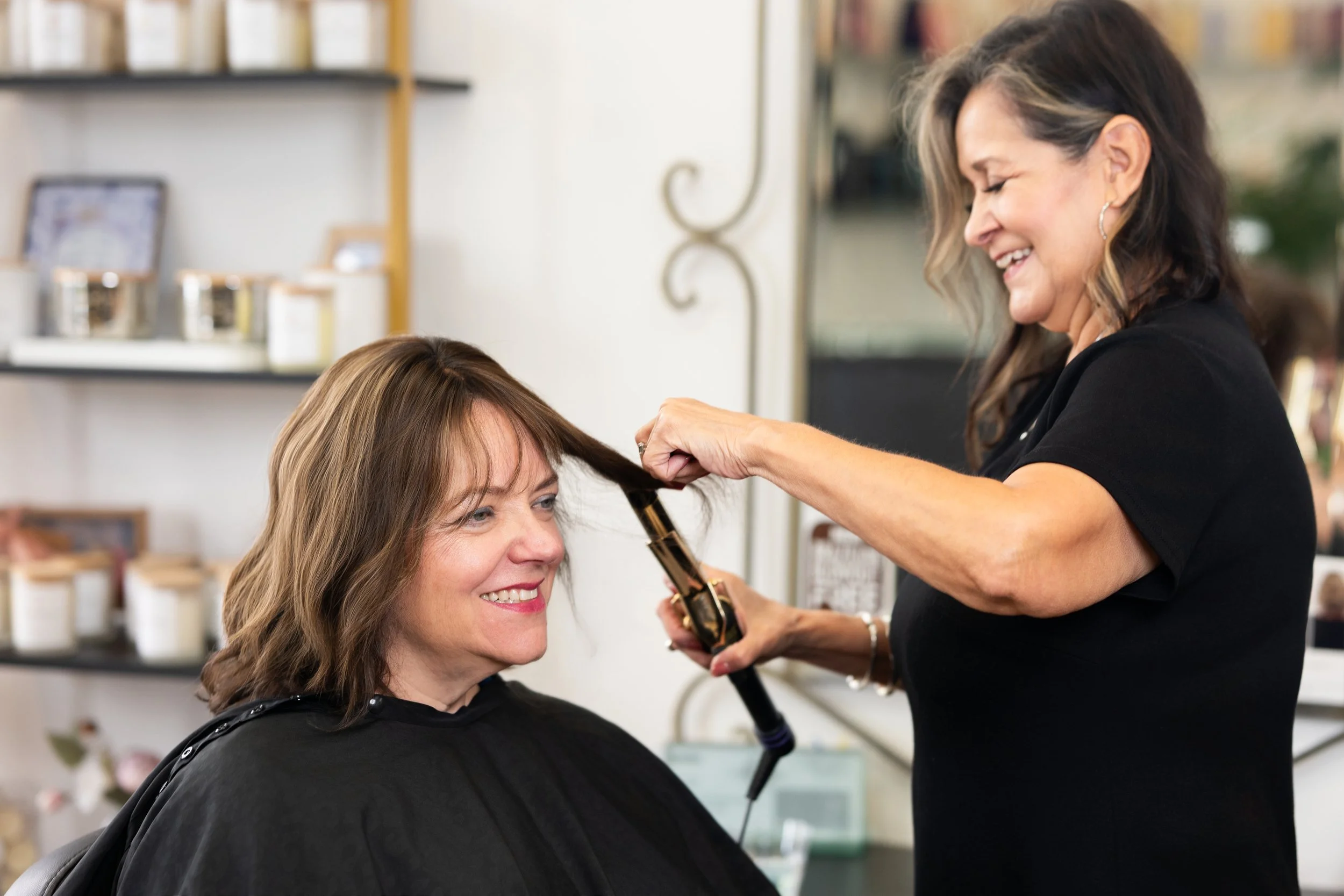 A woman with shoulder-length brown hair is smiling while having her hair styled by a hairstylist inside a salon.