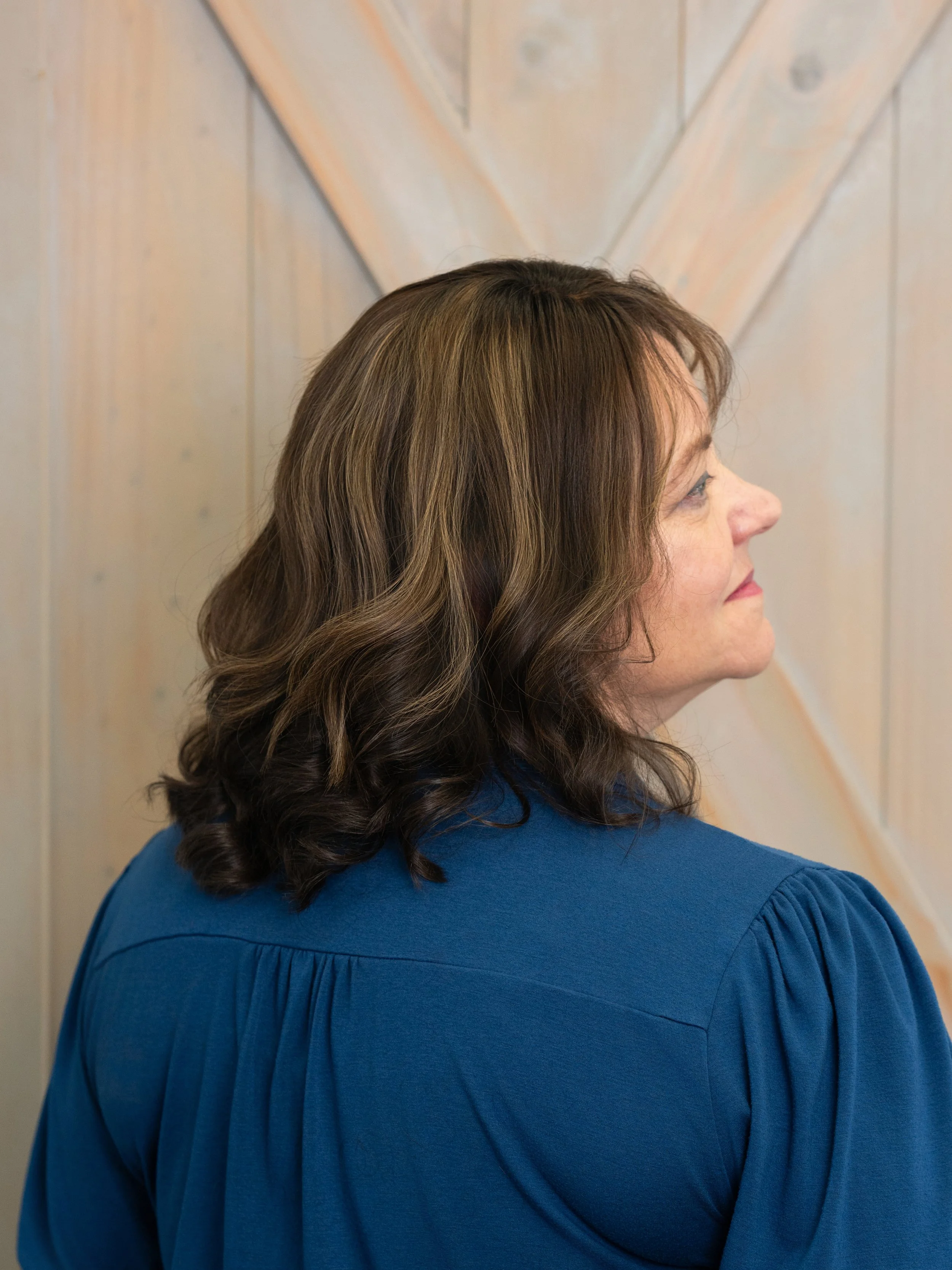 Profile side view of a woman with long wavy brown hair wearing a blue top, standing against a light-colored wooden wall.