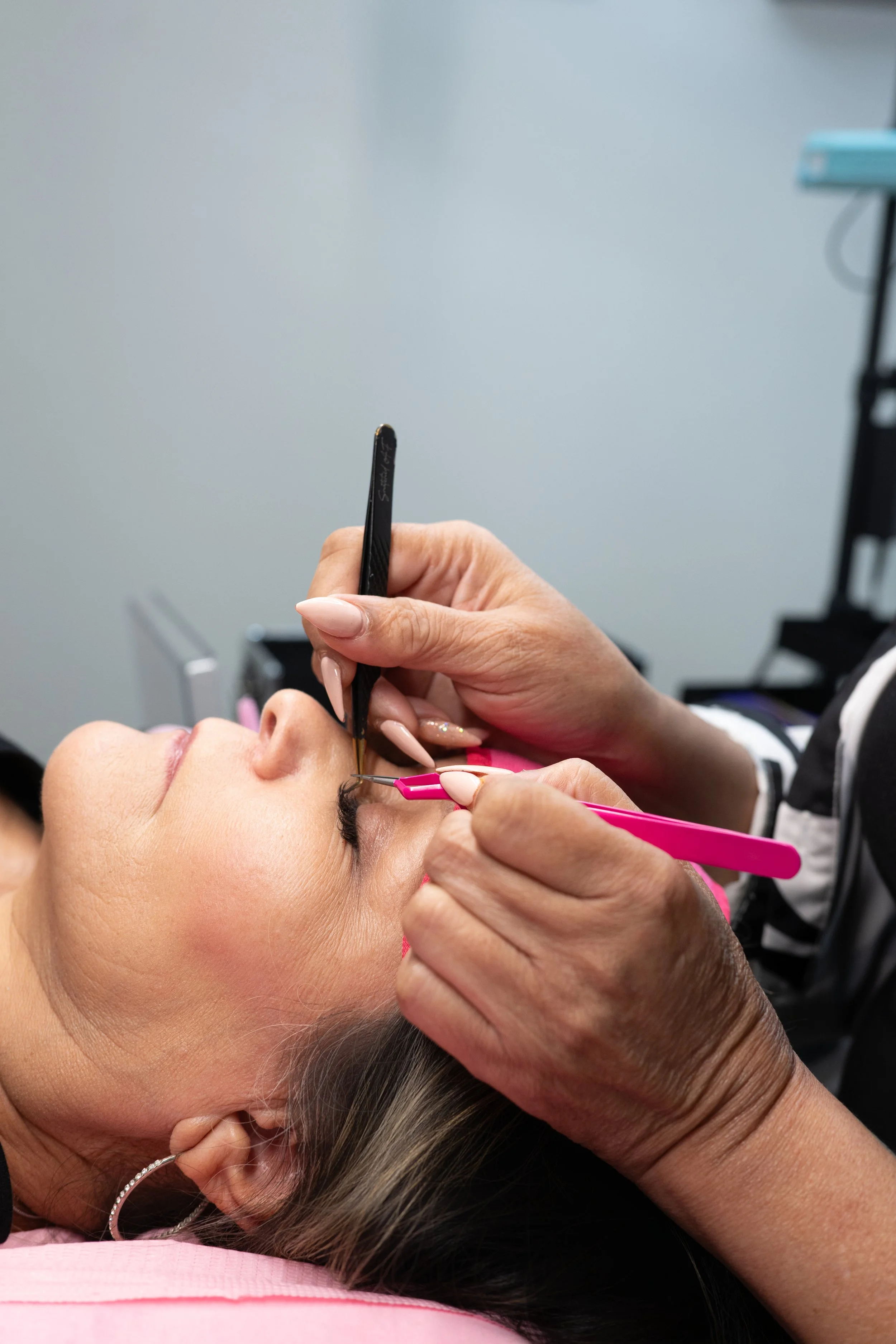 A woman receiving an eyelash extension treatment from a technician using specialized tools, lying down with eyes closed.