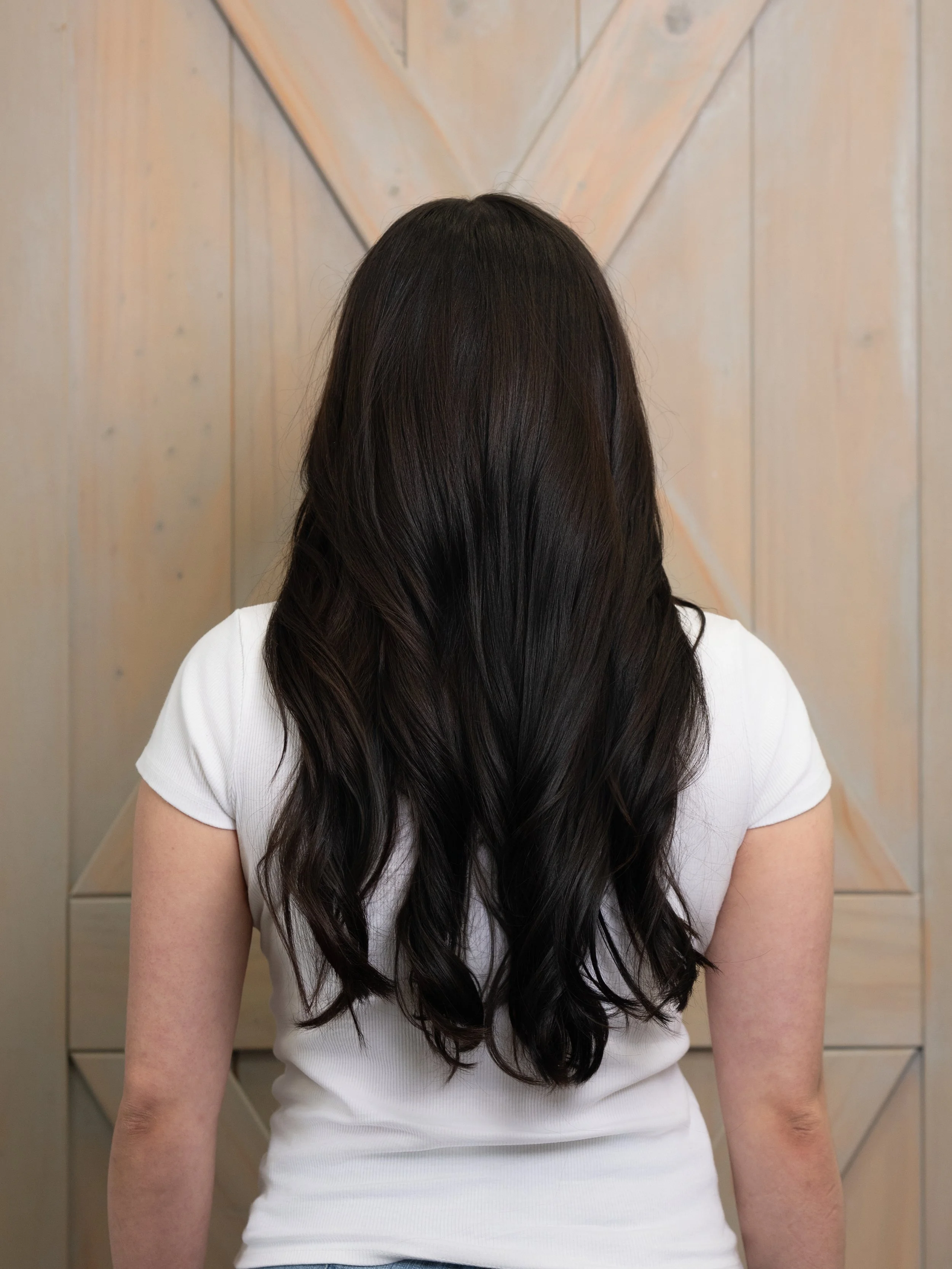 Back view of a woman with long, wavy dark hair, wearing a white shirt, with a wooden wall in the background.